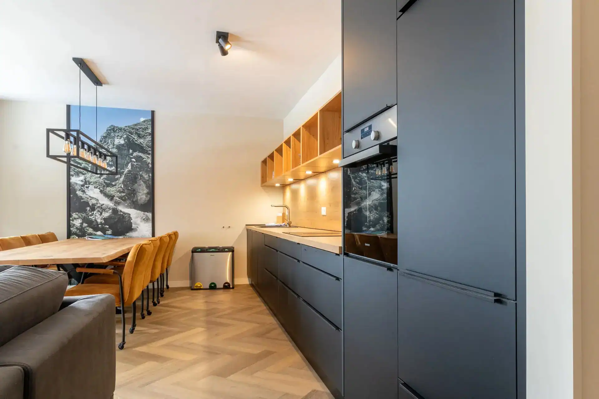 Detailansicht der modernen Einbauküche in den Silvretta Panorama Suites mit dunkelgrauen Fronten, warmen Holzelementen, angrenzendem Essbereich und einem großformatigen Landschaftsbild an der Wand.