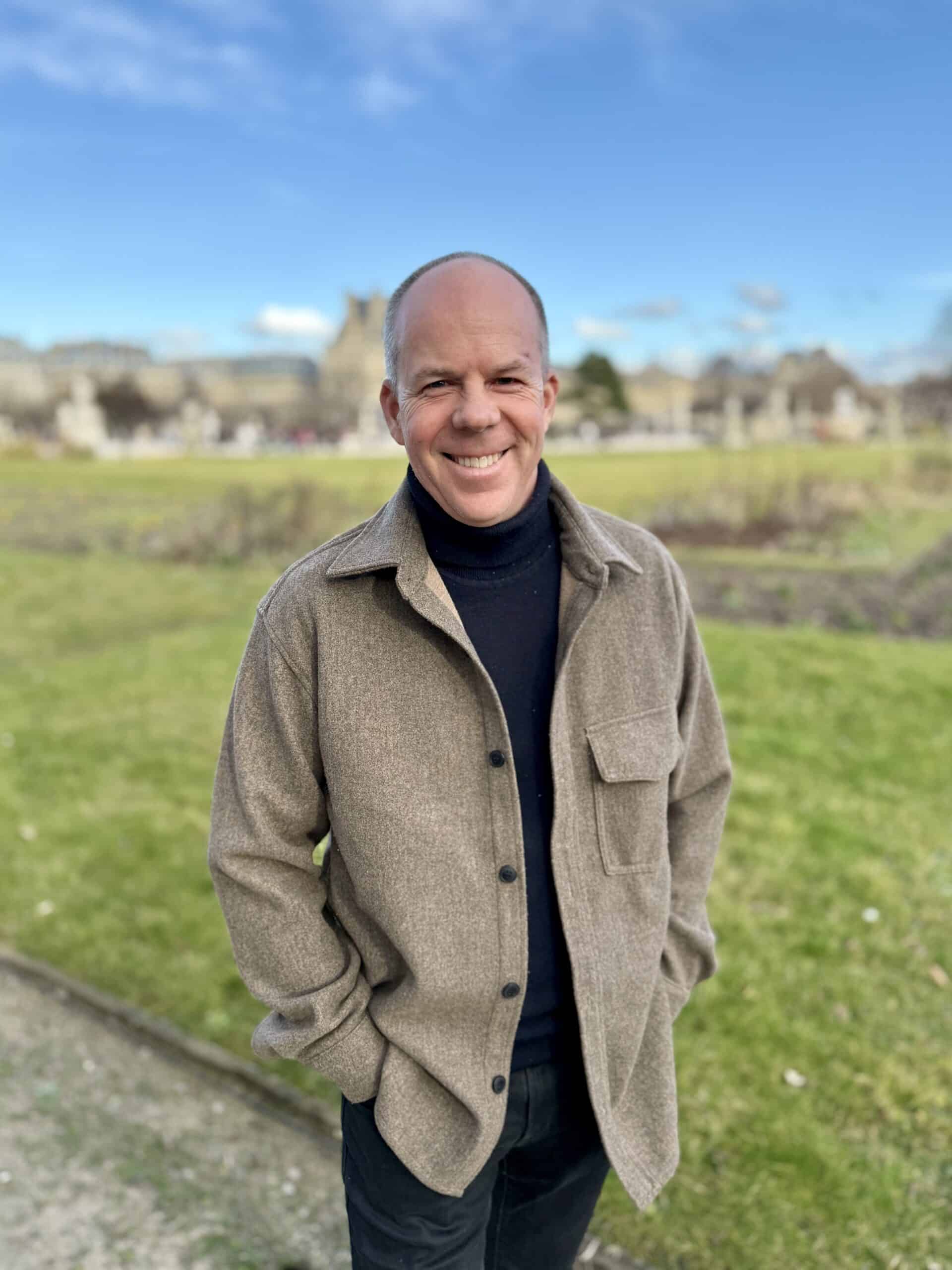 Derek Fredrickson, founder and CEO of The COO Solution standing in a field and smiling wearing a beige trench coat and a black turtleneck sweater