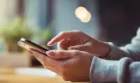 Mãos de uma pessoa usando um smartphone preto, com o dedo indicador tocando a tela, sobre uma mesa de madeira e fundo desfocado com luzes.