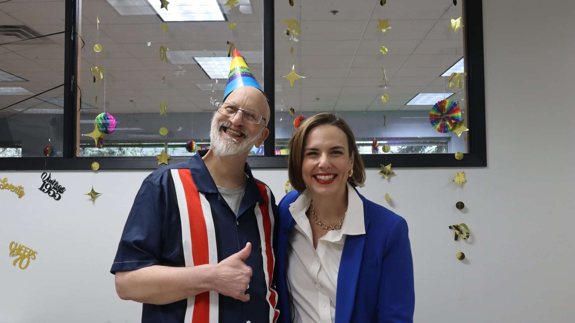 A man in a festive hat and striped shirt gives a thumbs-up while posing with a woman in a blue jacket. They are smiling in a decorated room with hanging stars and colorful ornaments.