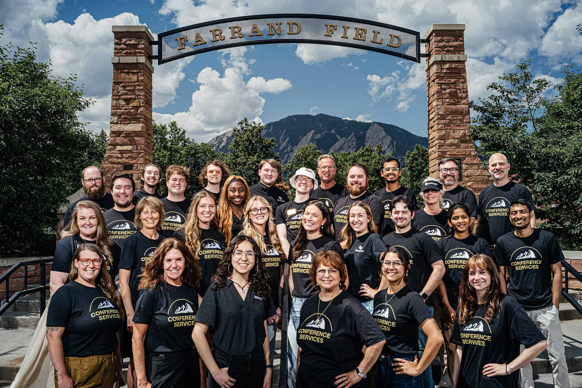 Group of conference services staff in black t-shirts with "Conference Services" text, posing in front of Farrand Field sign and mountains, promoting event management solutions for universities.