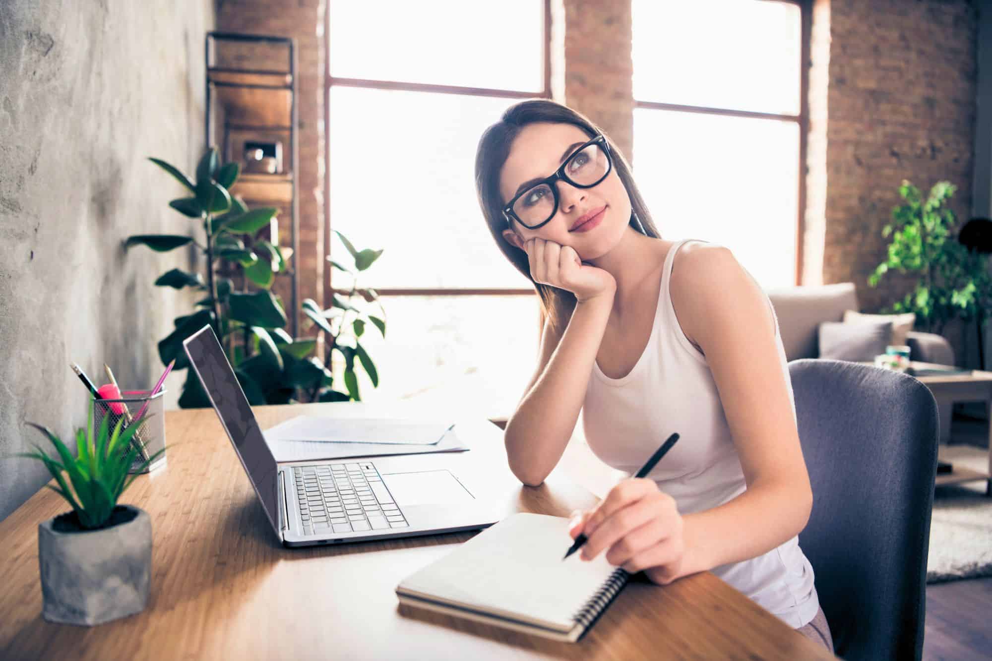 A single woman is at home, sitting at her desk, and looking off to think while making notes in her journal as she takes time to get ready to date.