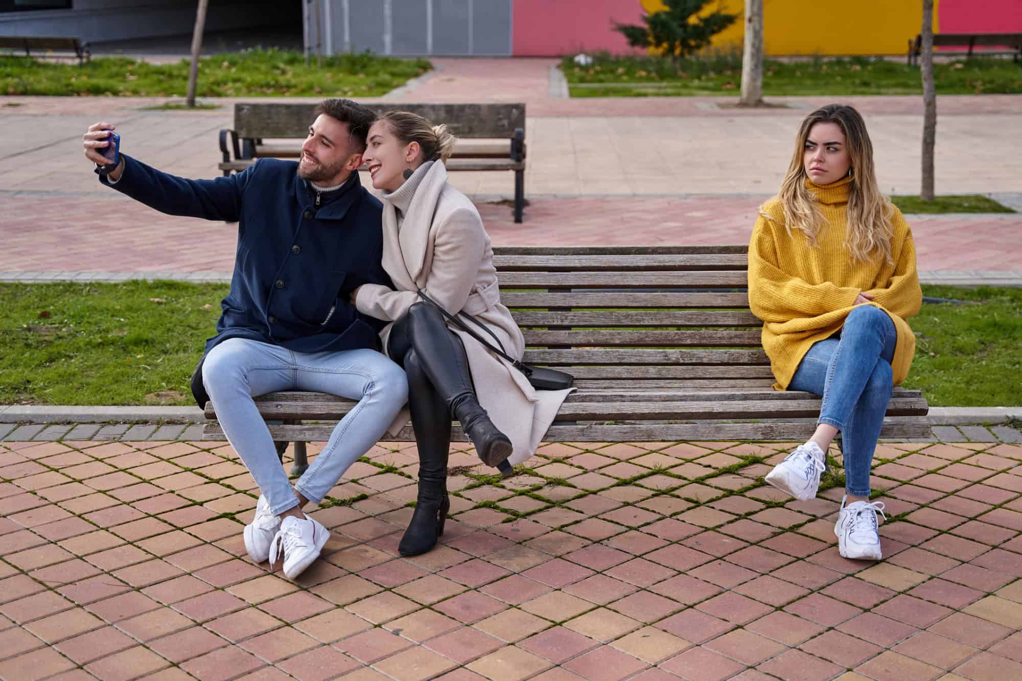 Loving couple taking a selfie while another woman jealously looks on.