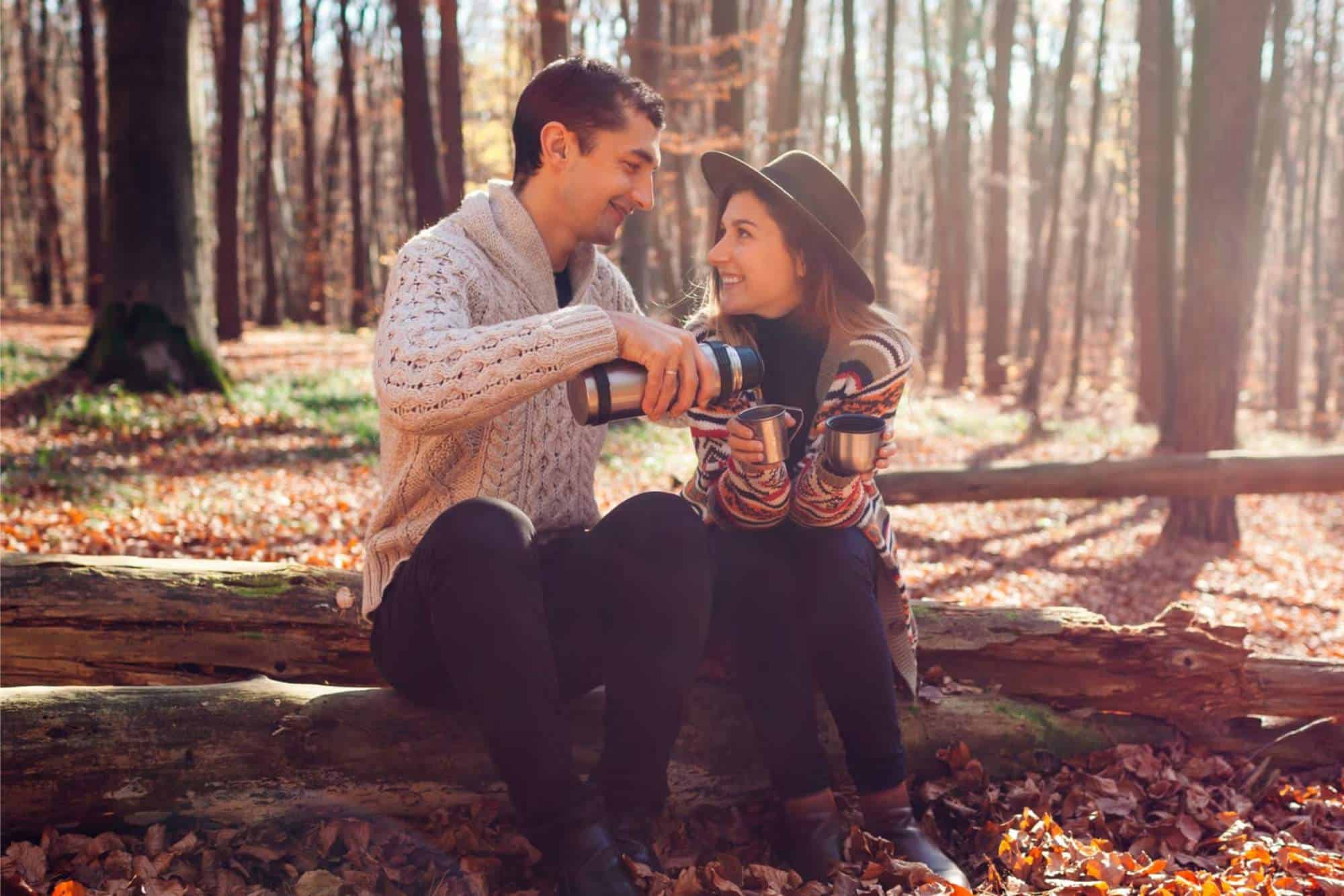 A couple sharing coffee from a thermos in the woods as they explore fall date ideas.