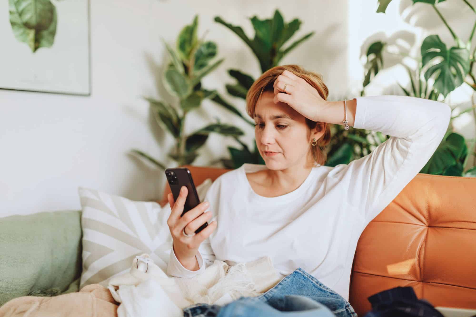 An attractive middle-aged woman sits on her couch and looks frustratingly at her phone as she navigates dating with AI.