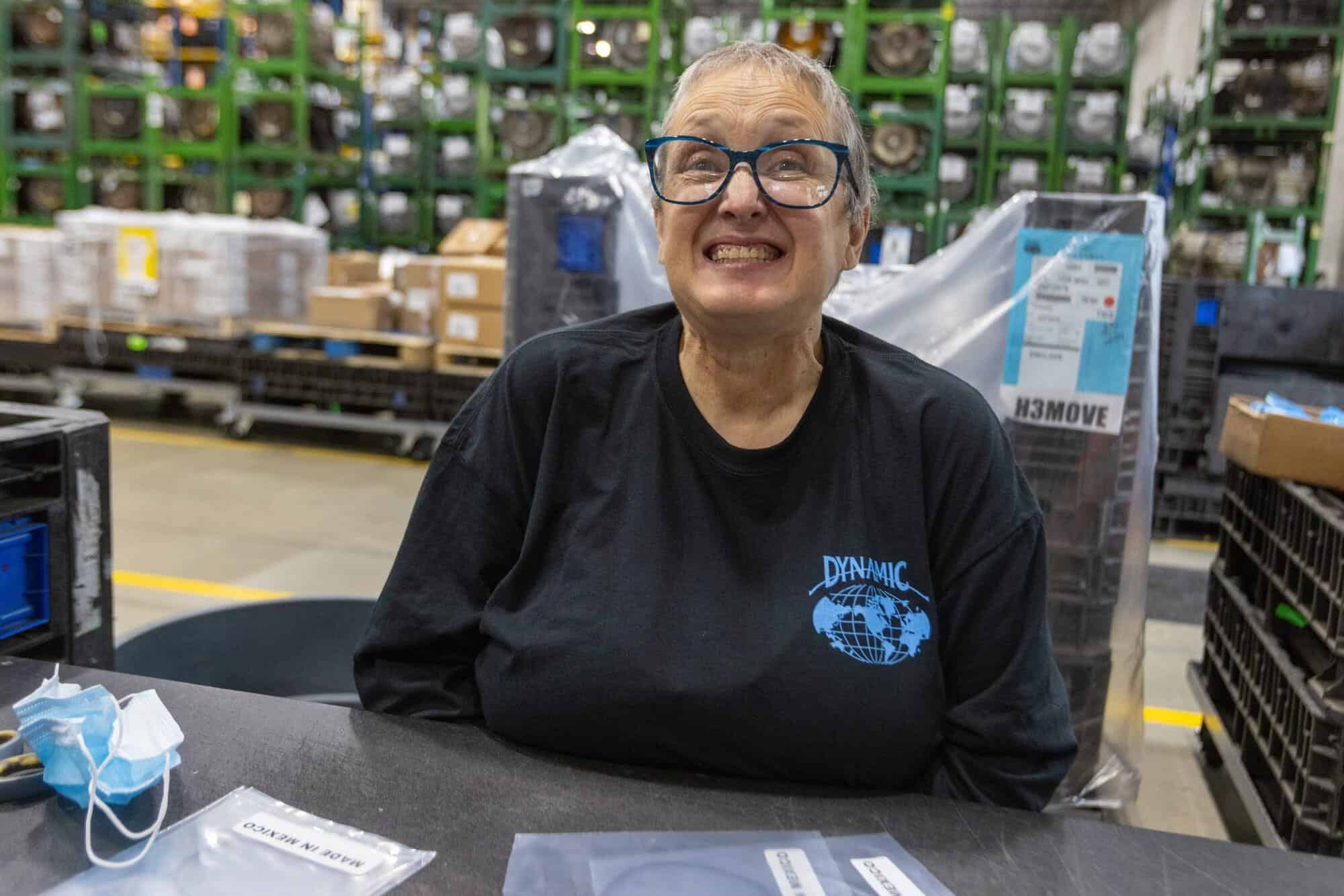 An older woman wearing glasses and a black shirt with a "Dynamic" logo smiles broadly in a warehouse setting. There's a table in front of her with packages and a face mask. Rows of shelves and bins are visible in the background.