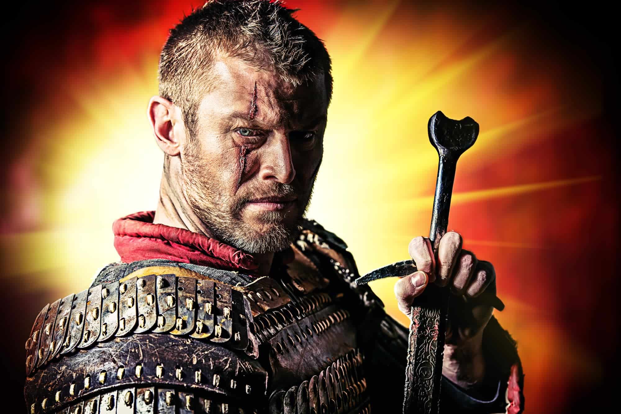 Fierce warrior with mud-streaked face, wearing leather armor and holding a sword with a cross-shaped handle. He stands against a fiery orange background with rays of light, creating a dramatic and intense atmosphere.