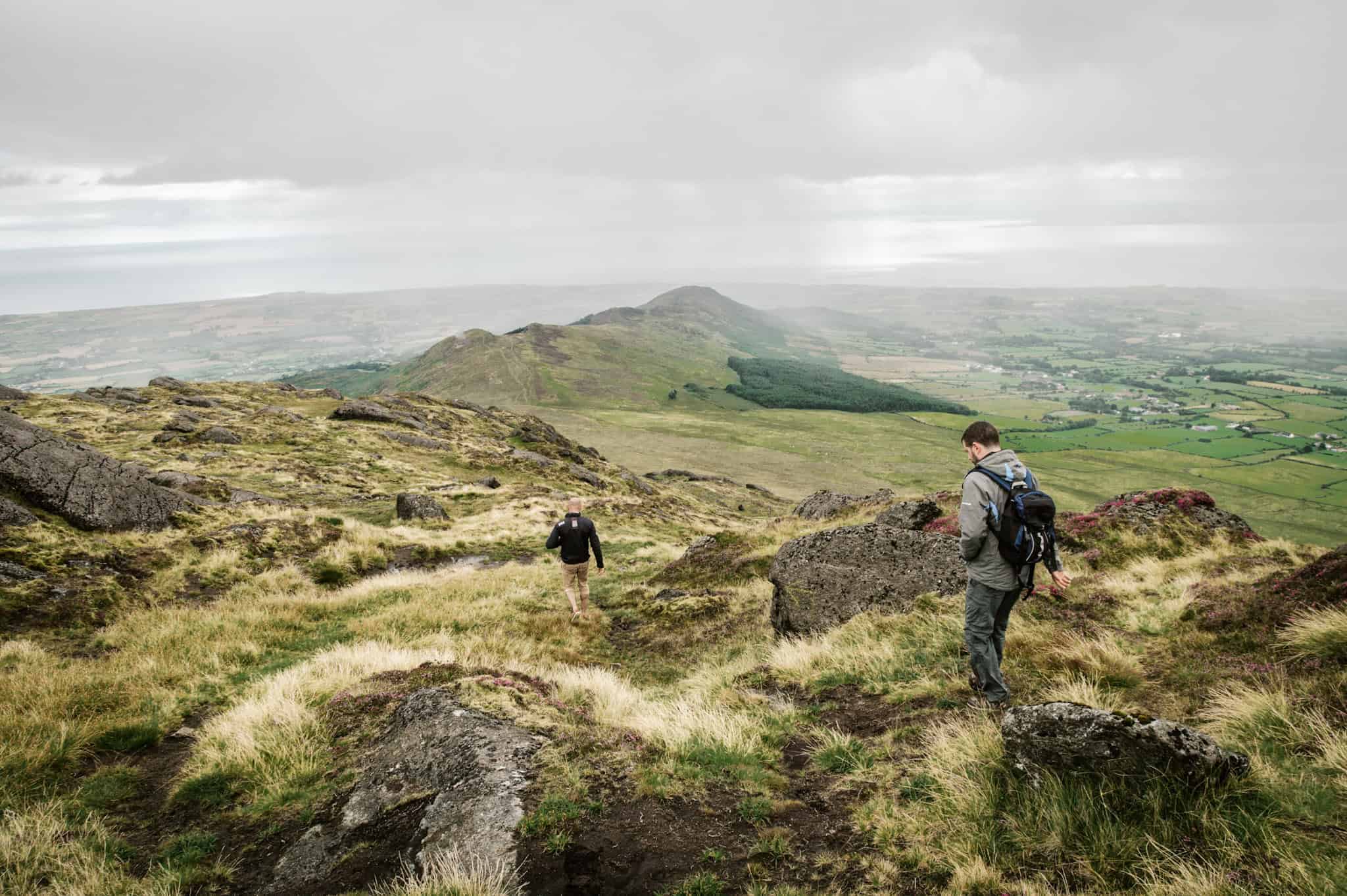 Two hikers walk across a grassy, rocky mountain slope with a misty view of Carlingford below. Rolling hills stretch into the distance under an overcast sky.