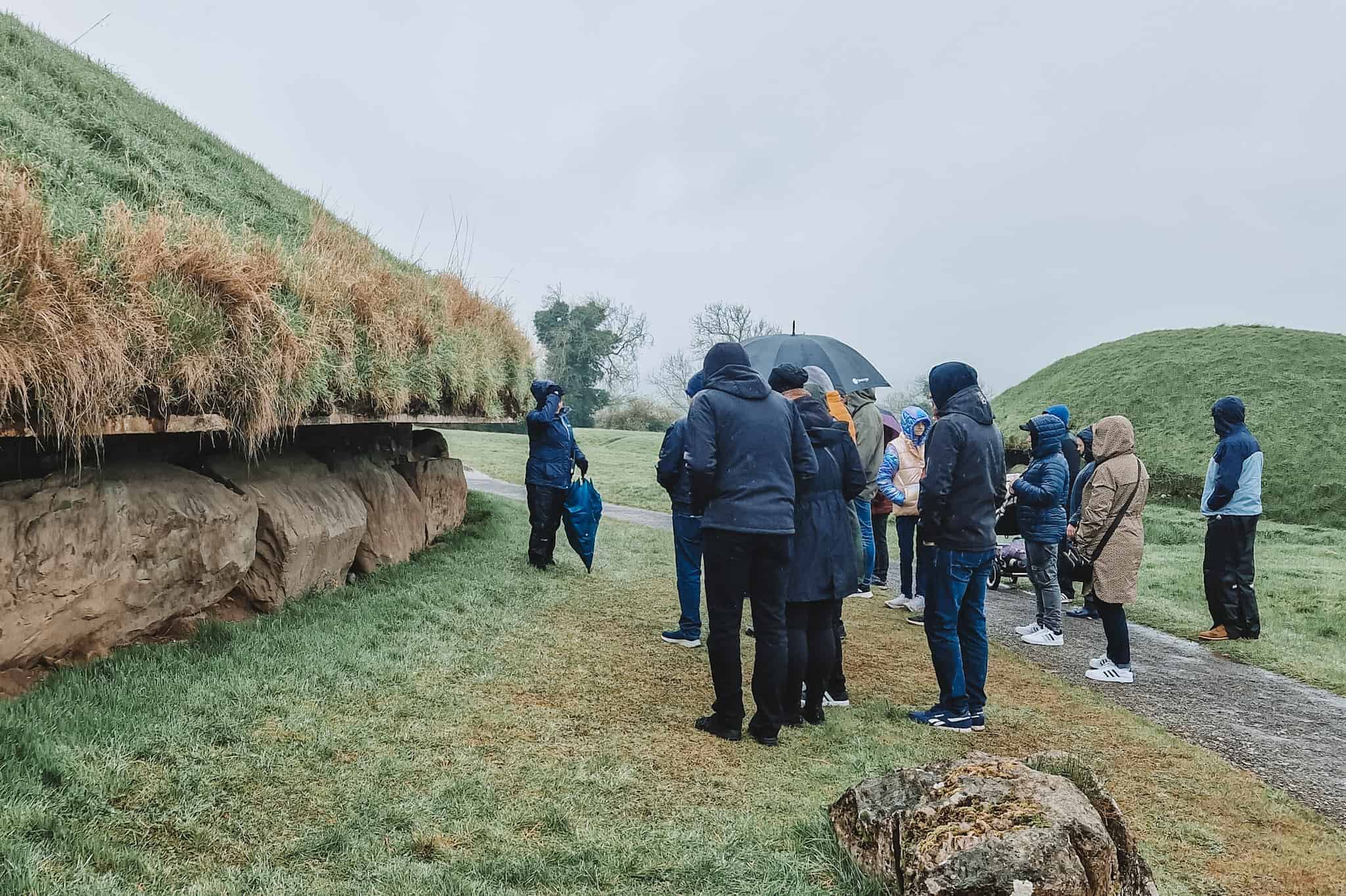 A group of tourists bundled up in rain gear, listening to a guide at Knowth, an ancient passage tomb in Ireland, on a wet and misty day.