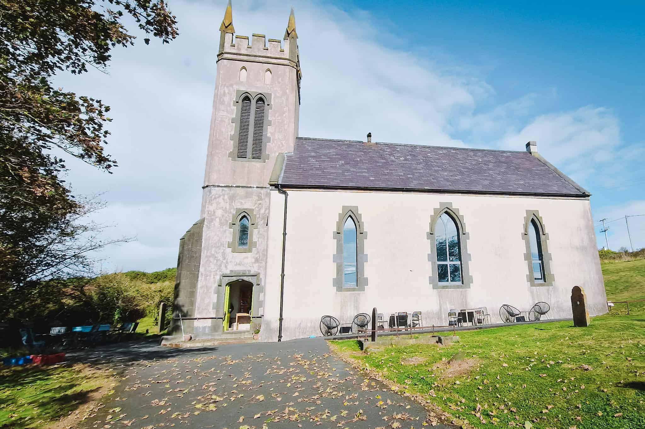 A small church with Gothic-style windows and a tall steeple, converted into a community space. The surrounding area includes a grassy lawn and scattered outdoor seating under a sunny sky.