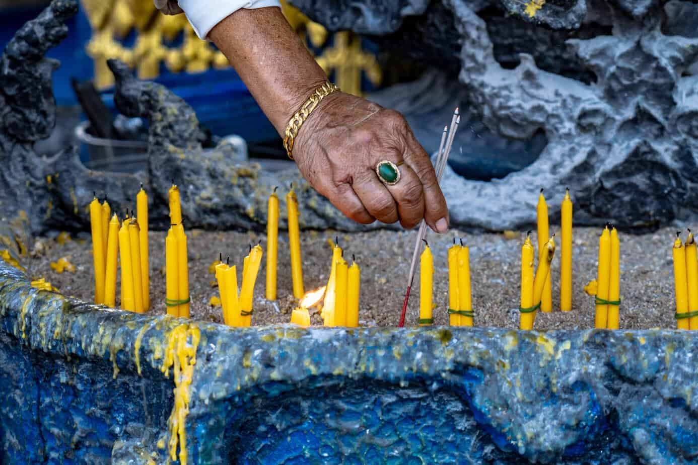 Chiang Rai: White Temple, Geheimtipps & Highlights im Norden Thailands 1 Eine Hand zündet Räucherstäbchen zwischen Reihen gelber Kerzen an, die in einem sandblauen Steinbehälter stehen.