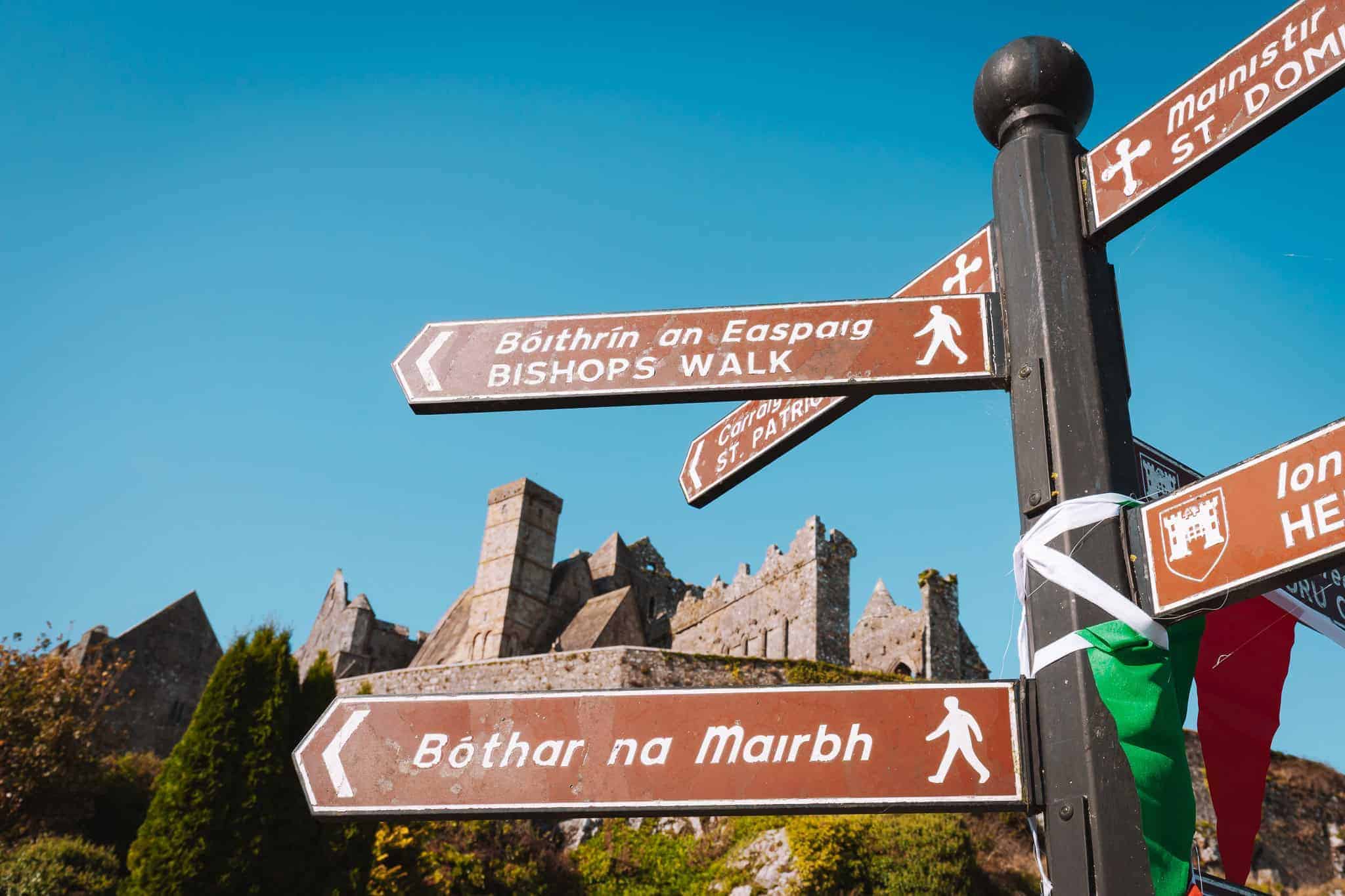 a close-up of a directional signpost in both Irish and English. The signs point toward locations such as "Bishop's Walk" (Bóithrín an Easpaig) and "Bóthar na Marbh" (Road of the Dead). In the background, the ruins of the historic Rock of Cashel rise against a bright blue sky, with stone structures and a mix of greenery, giving a sense of the ancient and the scenic landscape.