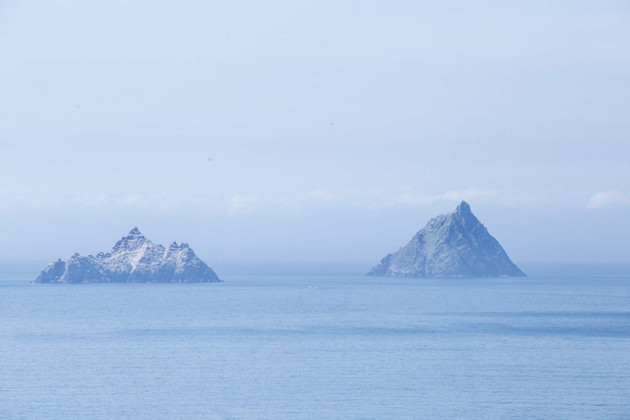 A serene view of the Skellig Islands off the coast of Ireland, rising sharply from the calm blue Atlantic Ocean. The larger island on the right, Skellig Michael, is covered in patches of green, while the smaller island on the left, Little Skellig, appears more rugged and rocky. The clear sky and soft haze create a peaceful and distant atmosphere.