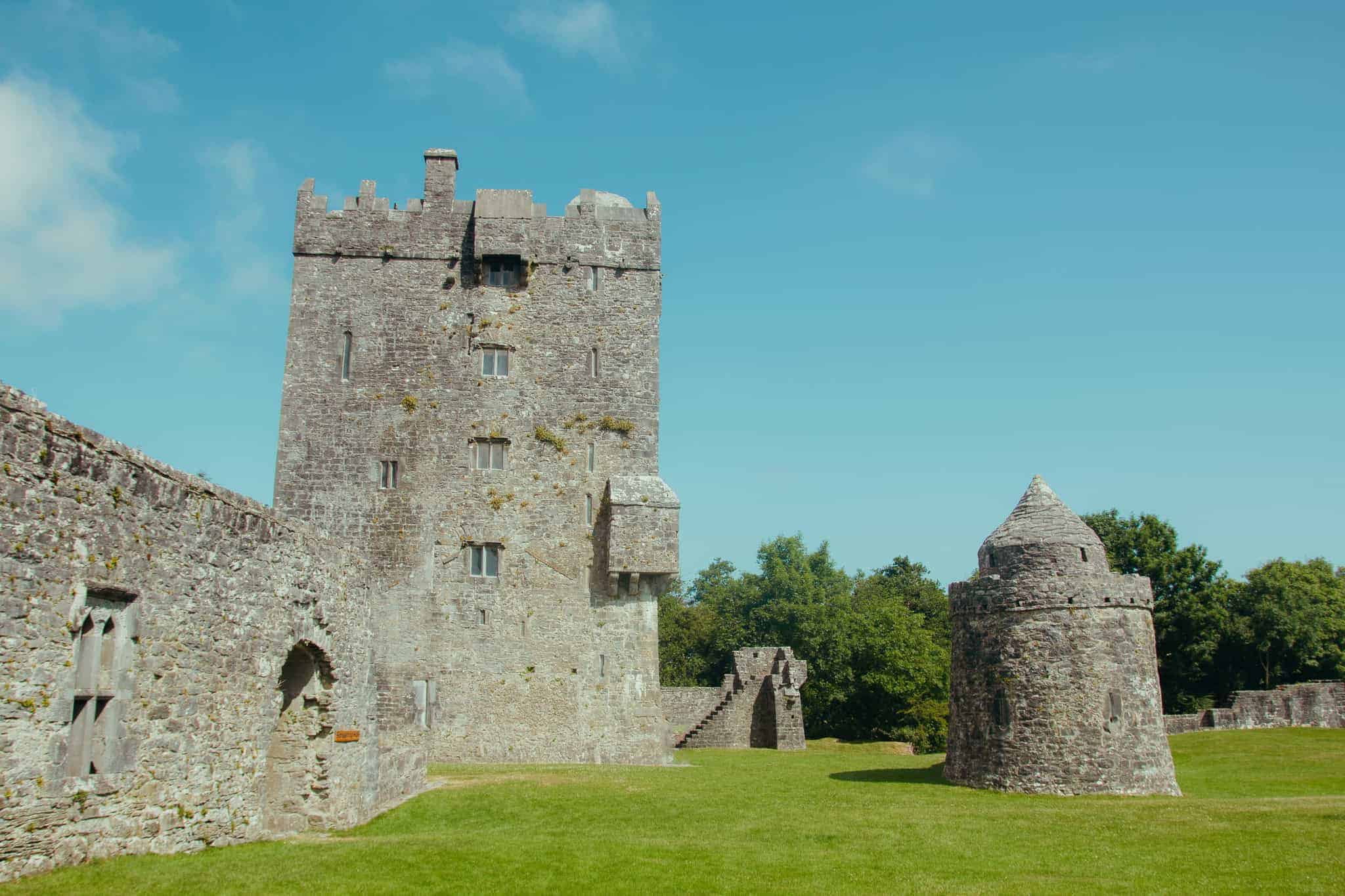 The historic stone structure of Aughnanure Castle in Ireland, featuring a tall tower and cylindrical outbuilding, surrounded by a well-kept lawn and trees under a clear blue sky.
