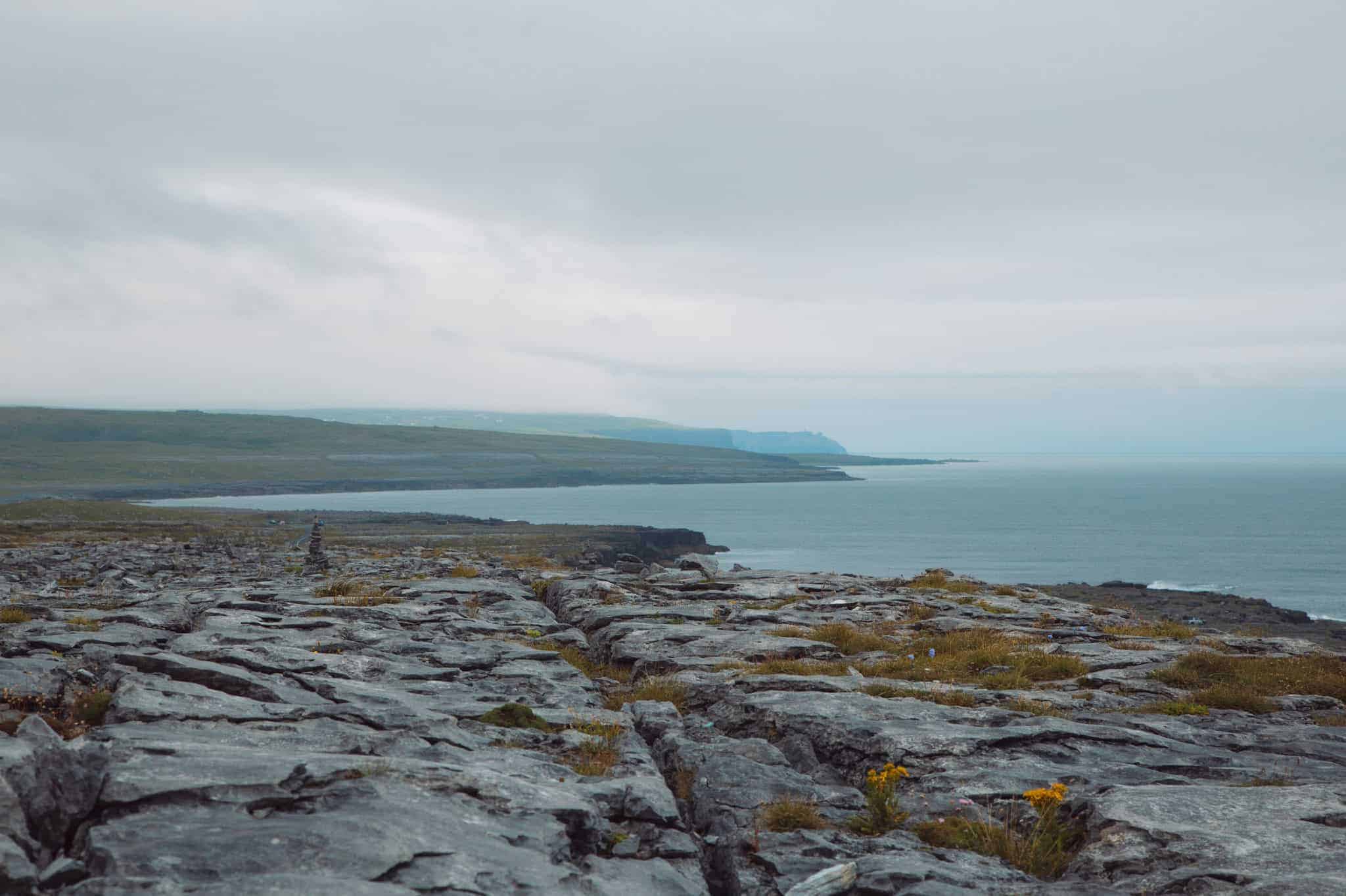 The rocky landscape of the Burren stretches towards the horizon, with the sea visible under a cloudy sky.