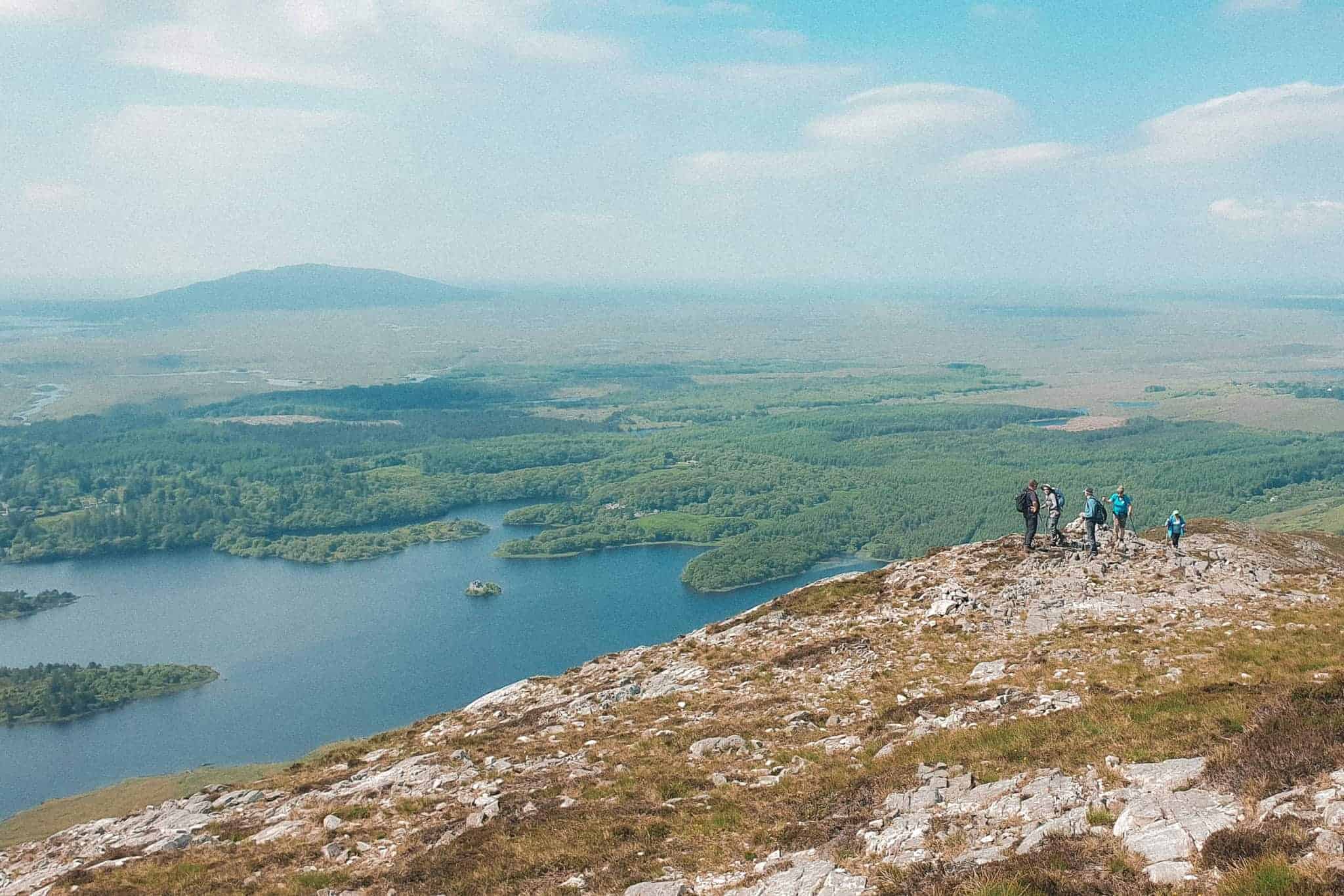 A group of hikers standing on a rocky outcrop, overlooking a vast landscape of lakes, forests, and distant mountains under a clear blue sky.