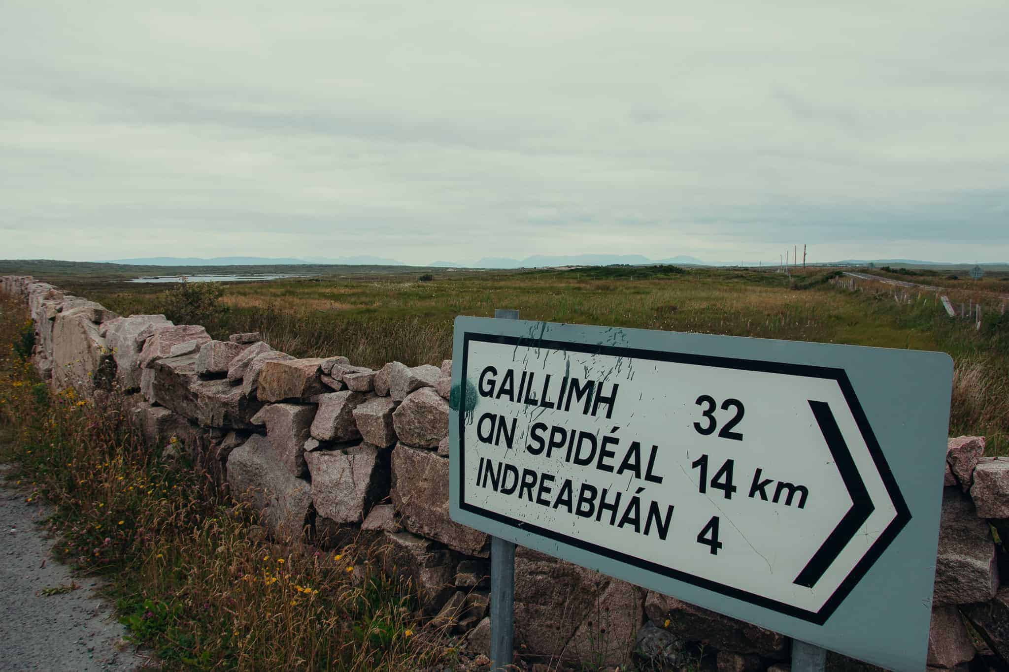 A road sign in the Irish countryside with directions in Gaelic: "Gaillimh 32 km," "An Spidéal 14 km," and "Indreabhán 4 km," set against a backdrop of stone walls and fields.