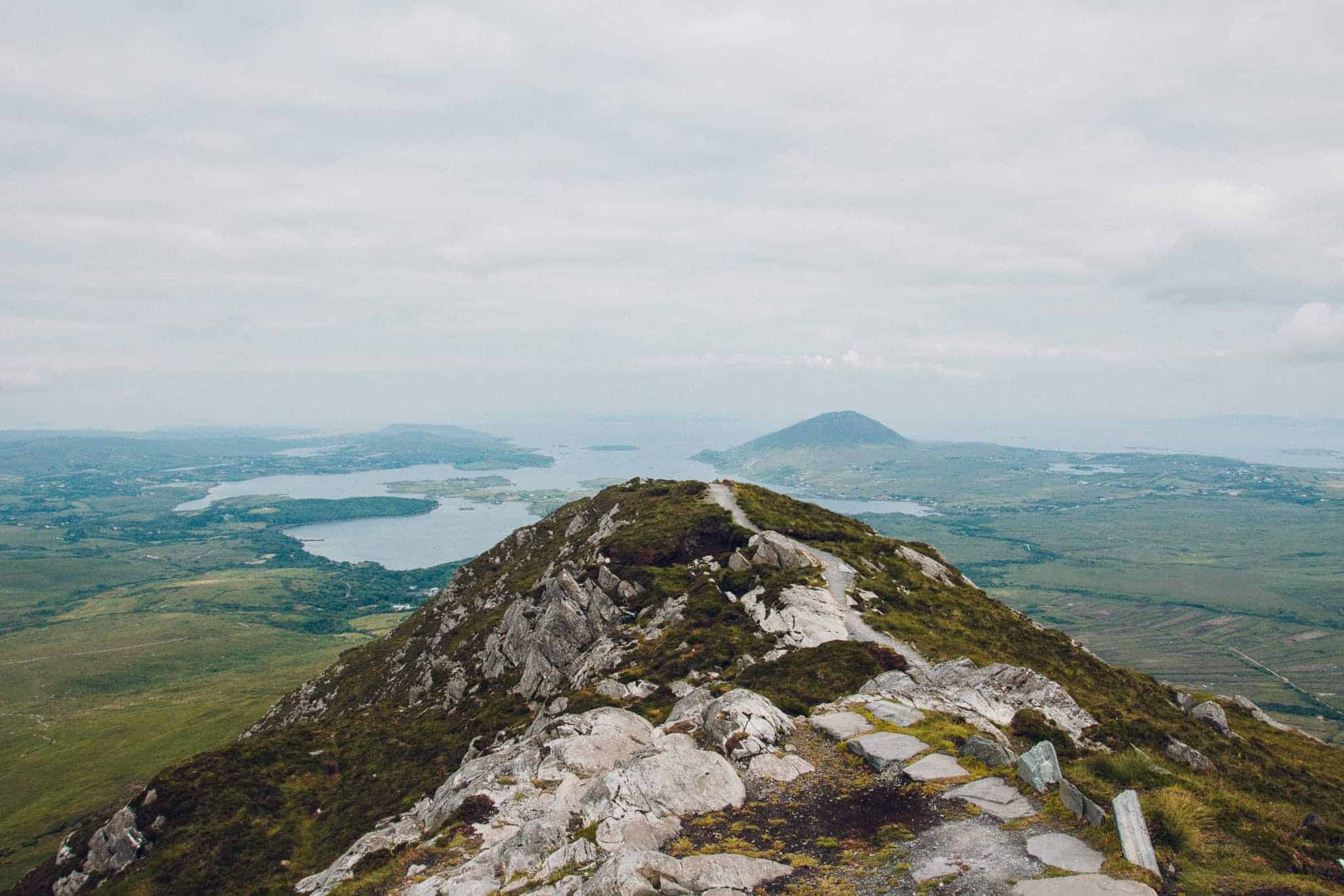 A rocky trail leading up to the peak of Diamond Hill, with expansive views of the surrounding green landscape and distant lakes under a cloudy sky.