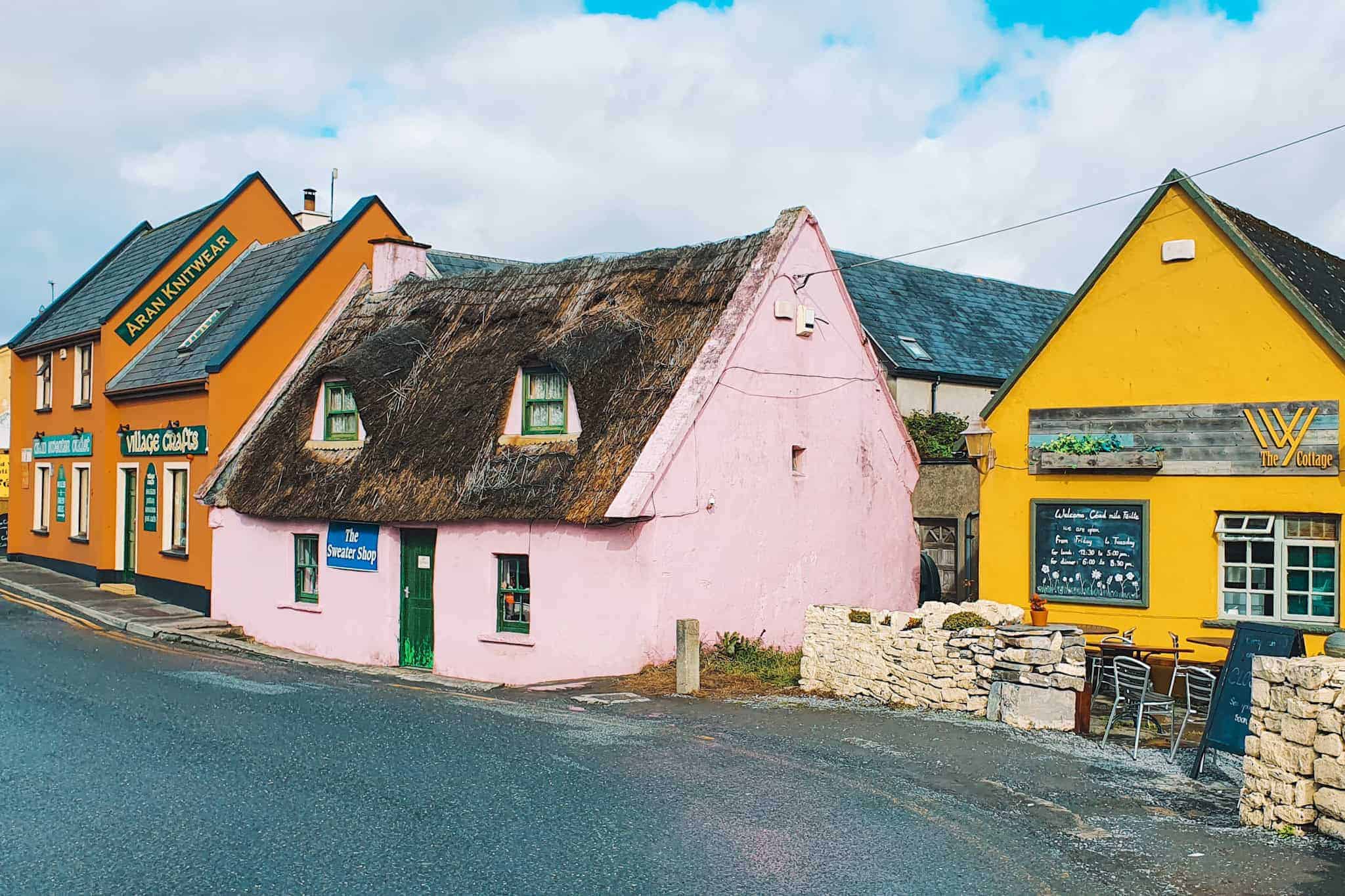 A quaint street in Doolin featuring a pink thatched-roof cottage labeled "The Sweater Shop," flanked by brightly colored buildings, including an orange one labeled "Aran Knitwear" and a yellow one labeled "The Cottage," under a partly cloudy sky.