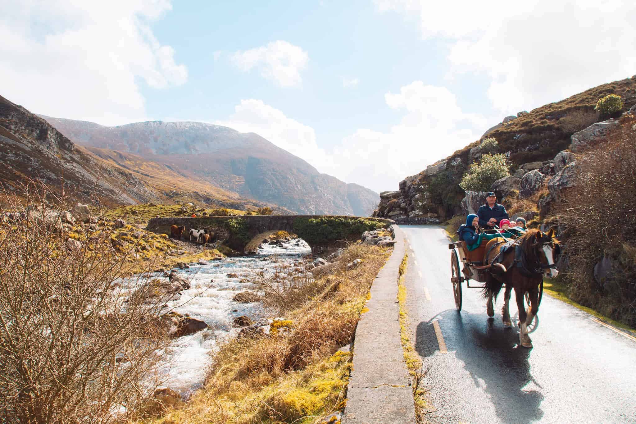 A horse-drawn carriage carrying people along a winding mountain road in the Gap of Dunloe, with a river and stone bridge in the background under a bright sky.