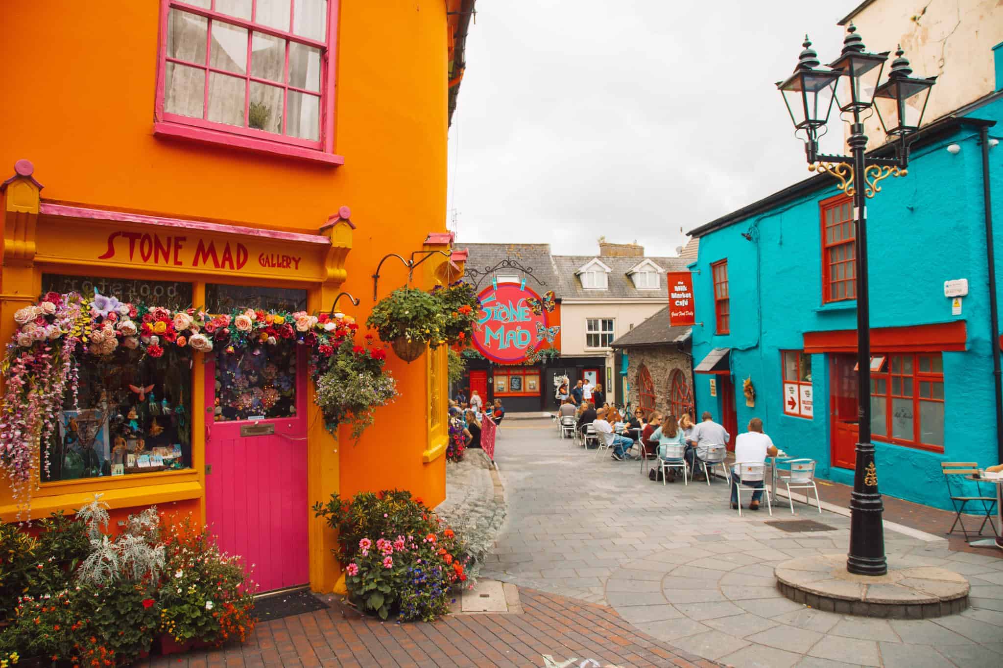 A vibrant, colorful street scene in Ireland featuring the "Stone Mad Gallery," a bright orange building with a hot pink door and window frames, adorned with hanging flower baskets and floral decorations. Across the cobblestone street is a turquoise blue building with red trim, where people are seated at outdoor café tables, enjoying a relaxed atmosphere. Classic black street lamps add charm to the lively, artistic setting.