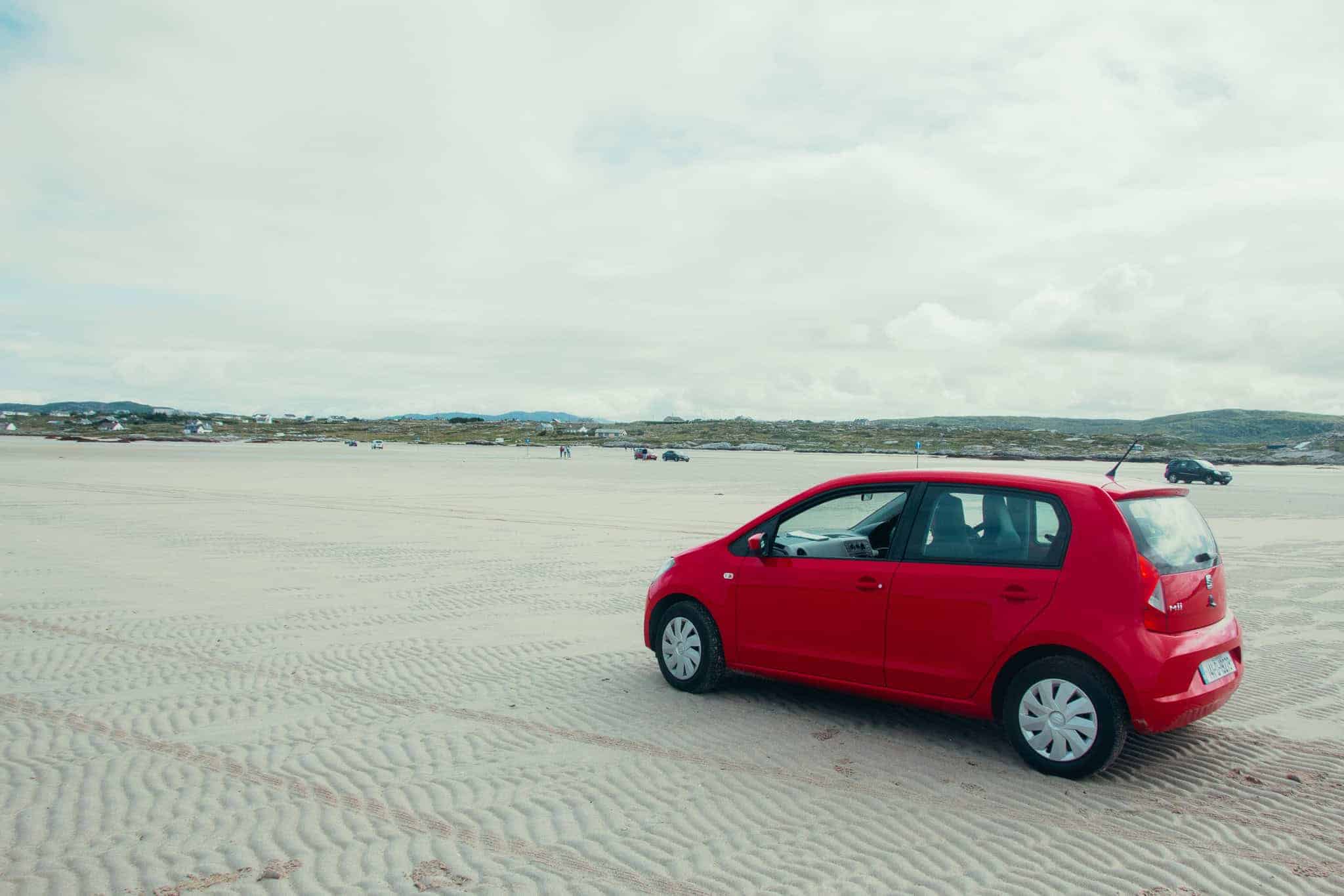 A red car driving to on a vast sandy beach to Omey Island, with distant hills and scattered houses in the background.
