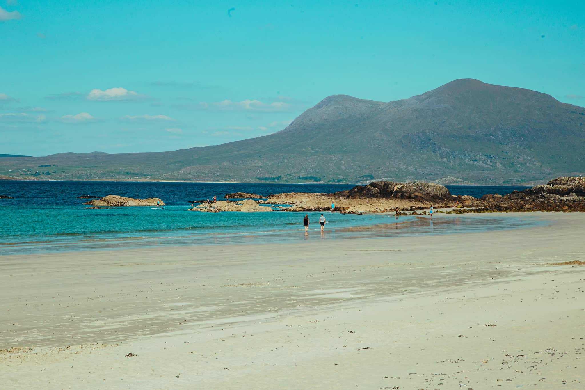 A sandy beach with turquoise waters and people walking along the shoreline, with mountains in the background.