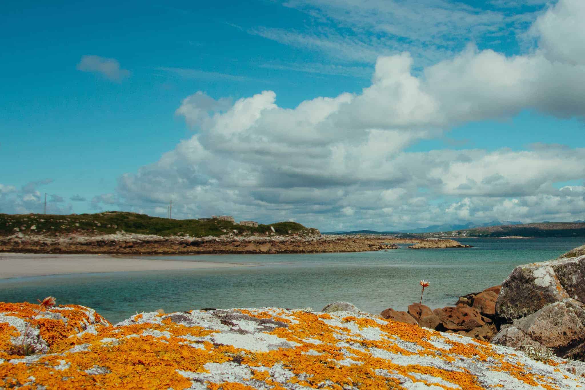 A beach with vibrant orange and white moss-covered rocks overlooking a calm, blue sea and distant hills.