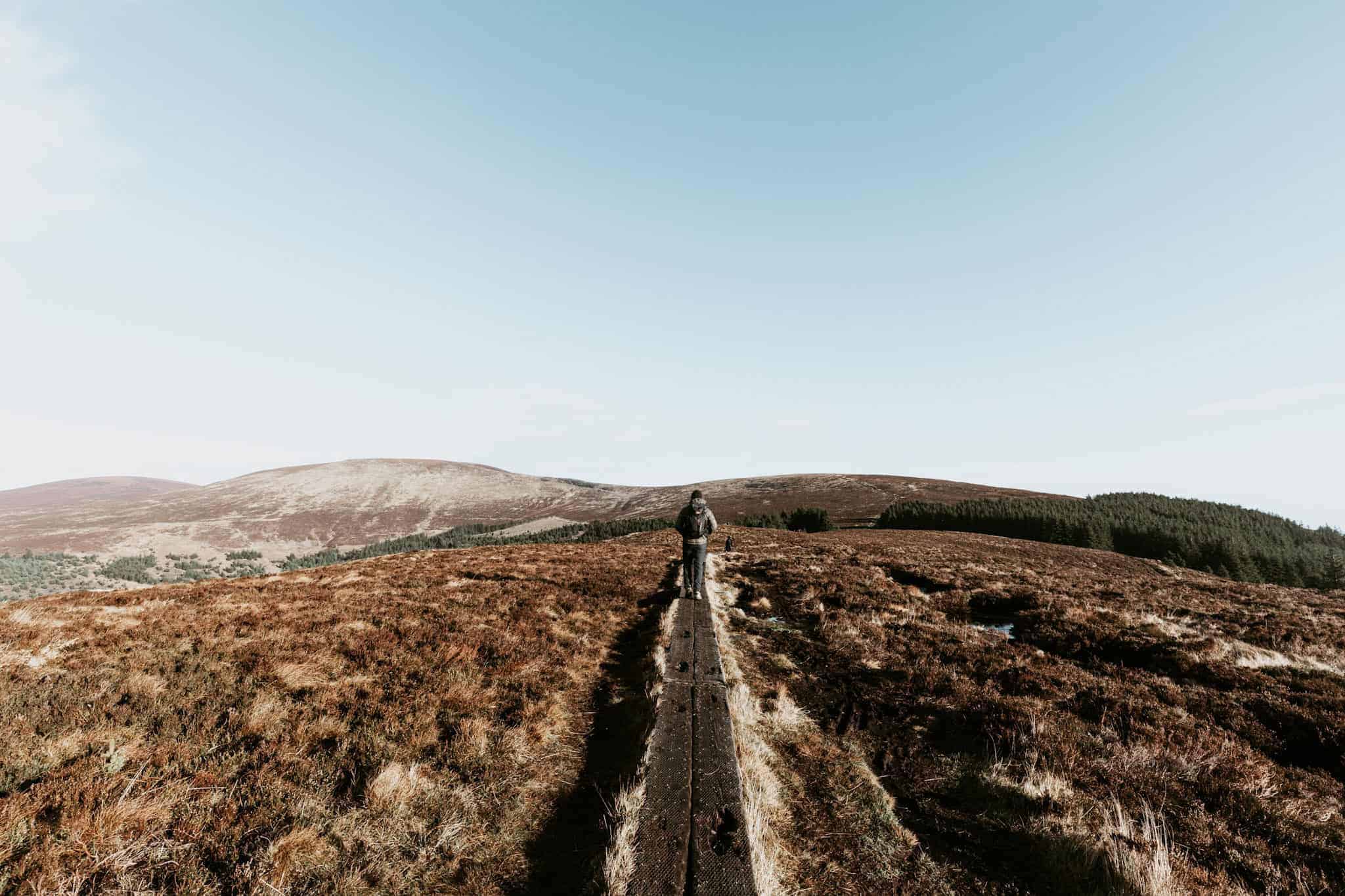 A lone hiker walks down a wooden boardwalk that stretches across a vast, brown moorland. The horizon is lined with gently sloping hills under a clear, pale blue sky.