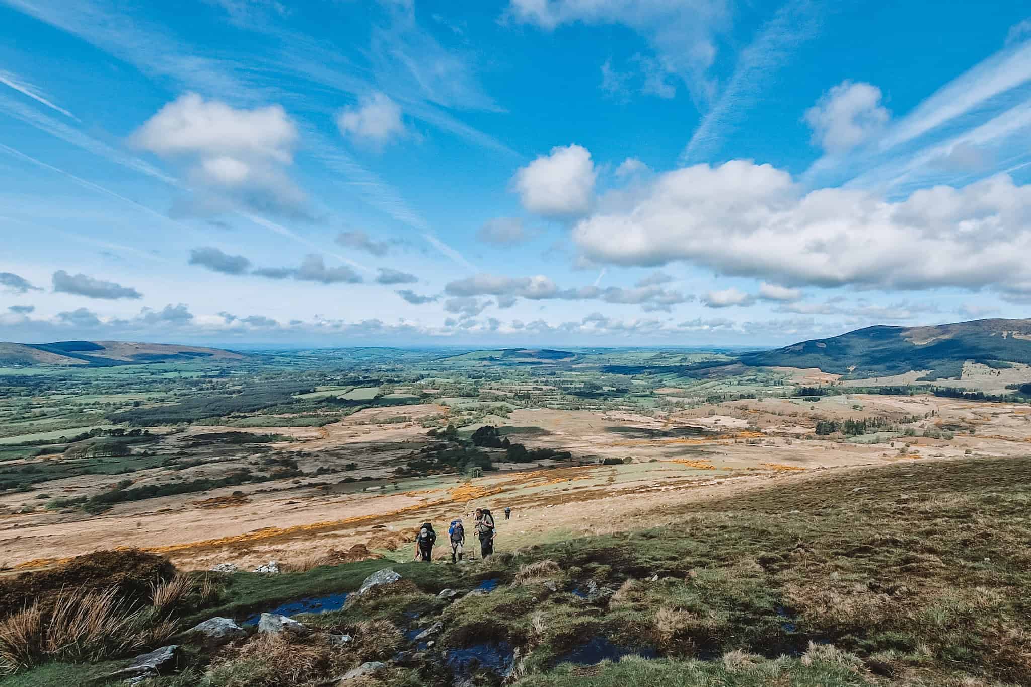 A wide view of a sprawling valley, with hikers walking up a grassy slope towards distant hills. The landscape is dotted with patches of yellow gorse and rocks under a bright, expansive sky.