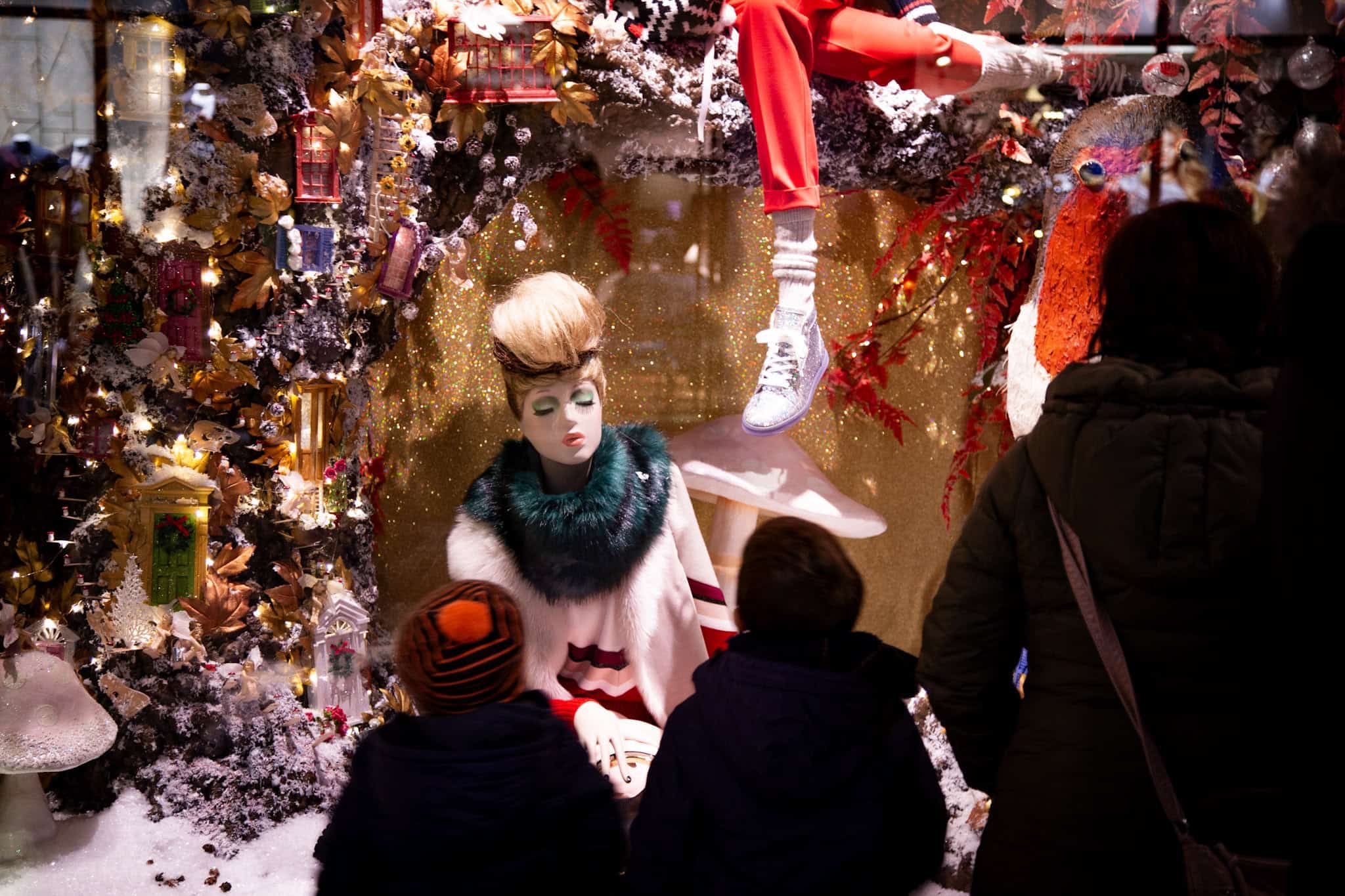 A festive store display featuring a mannequin in a luxurious winter outfit, surrounded by sparkling fairy lights, miniature decorative houses, and frosted foliage. Two children in the foreground gaze at the whimsical holiday scene.