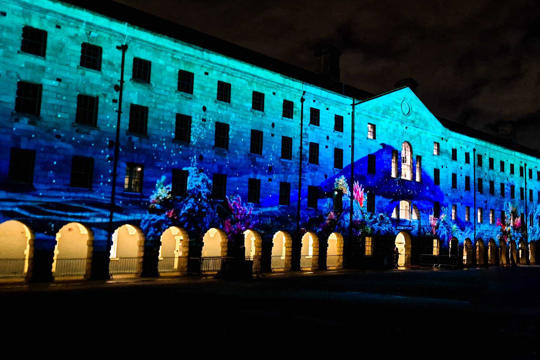 building facade lit with an underwater-themed projection featuring vibrant blue tones and coral reef imagery. Arched doorways at the base are warmly illuminated.