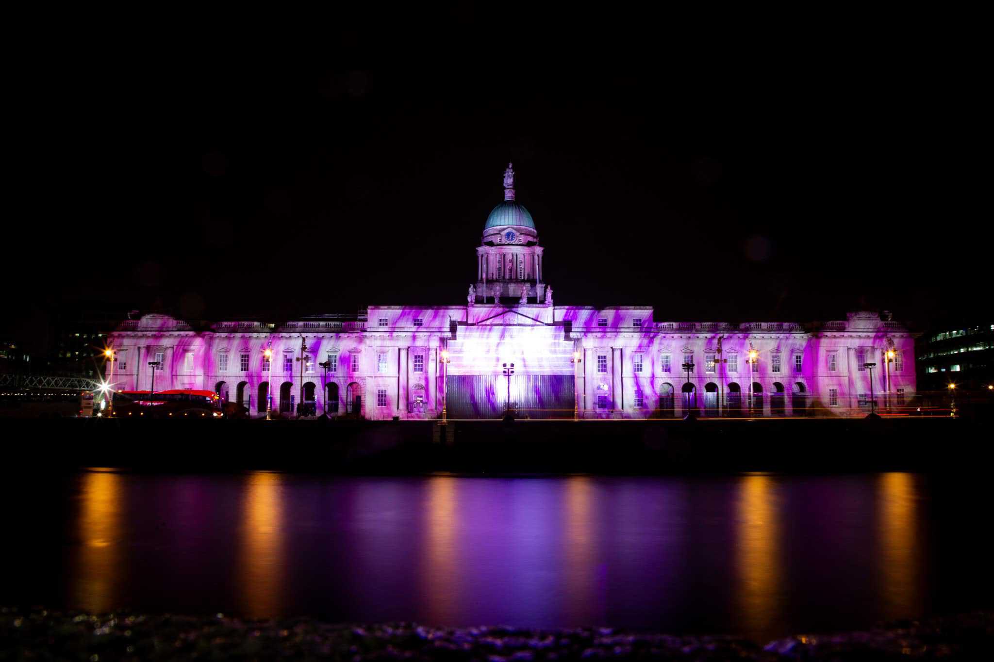 A historic building featuring a dome, lit in vibrant pink and purple tones, reflecting beautifully on the water below. The architectural details of the facade are accentuated by the colorful lighting.