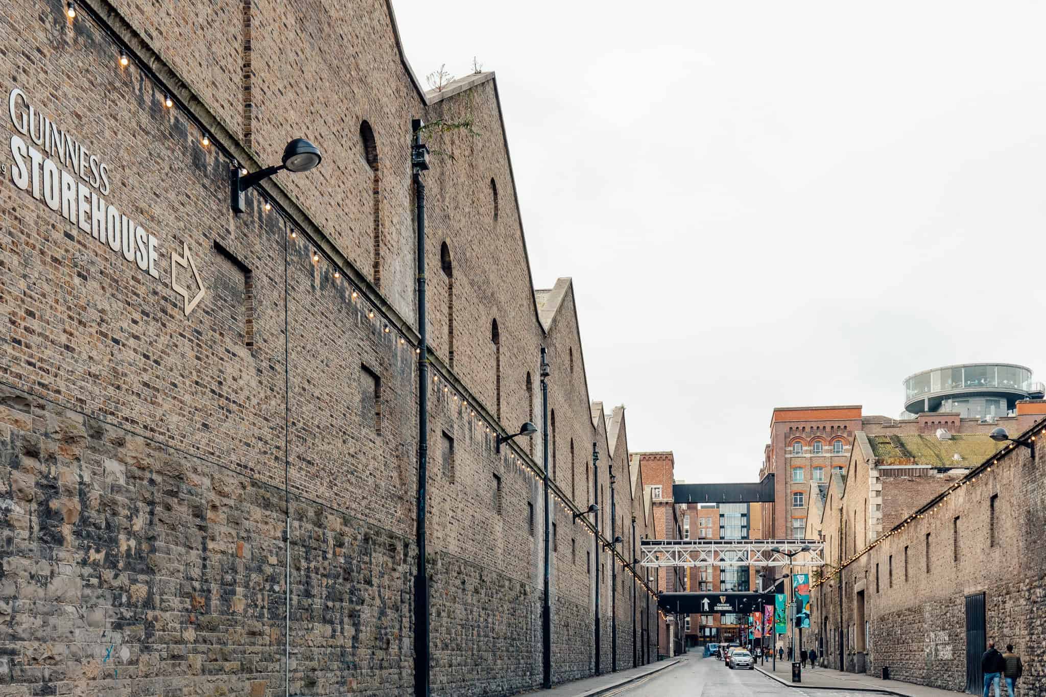 A view of the Guinness Storehouse, with its brick facade stretching along a narrow street lined with colorful banners and streetlamps. The backdrop includes modern and historic buildings, creating a blend of industrial heritage and urban scenery.