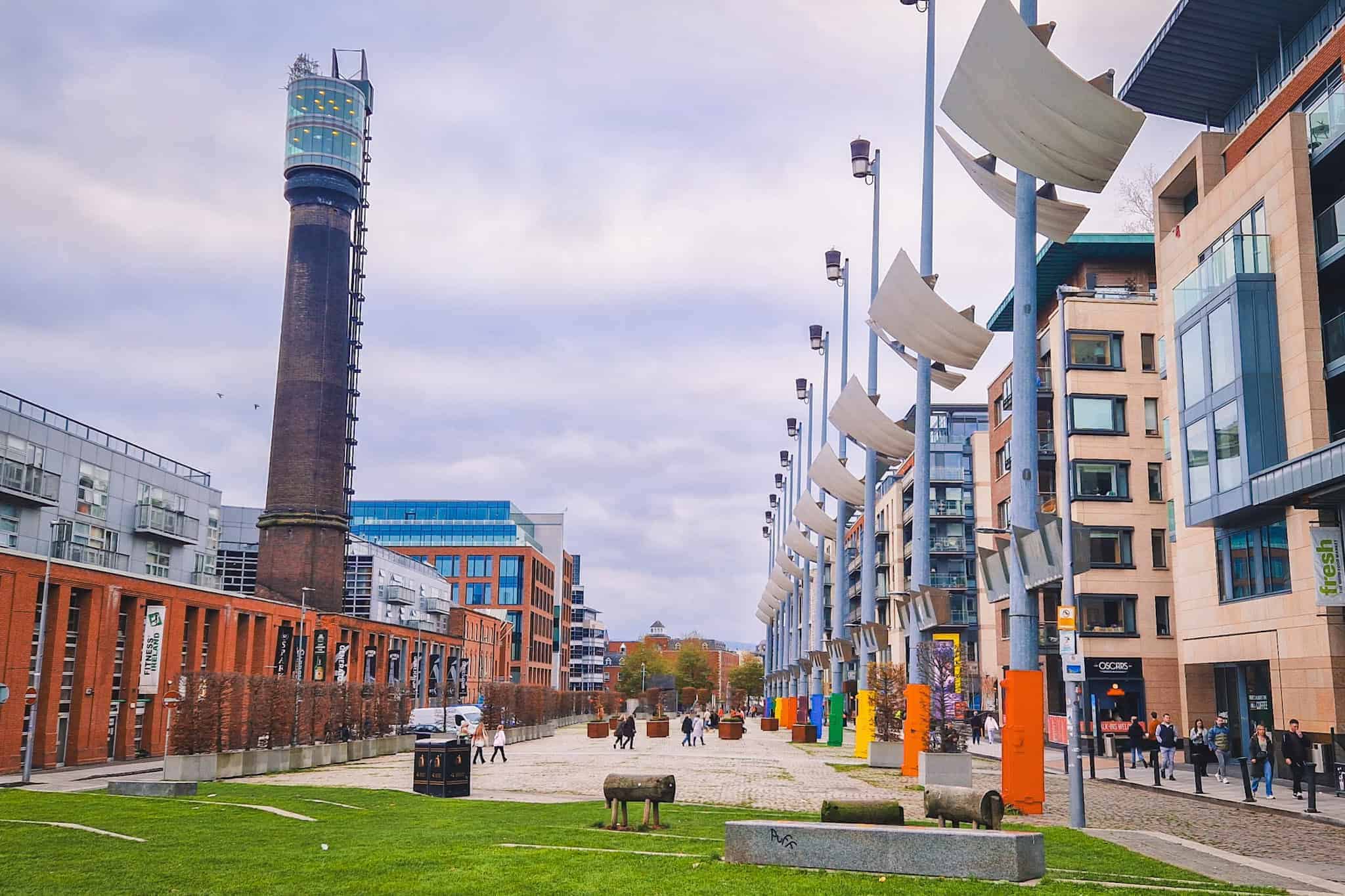 A wide view of Smithfield Square in Dublin, lined with modern buildings and distinctive tall lamp posts featuring sail-like panels. The scene includes a tall brick tower with an observation deck on the left and scattered benches and decorative elements in the square. People walk along the open space, adding life to the urban landscape.