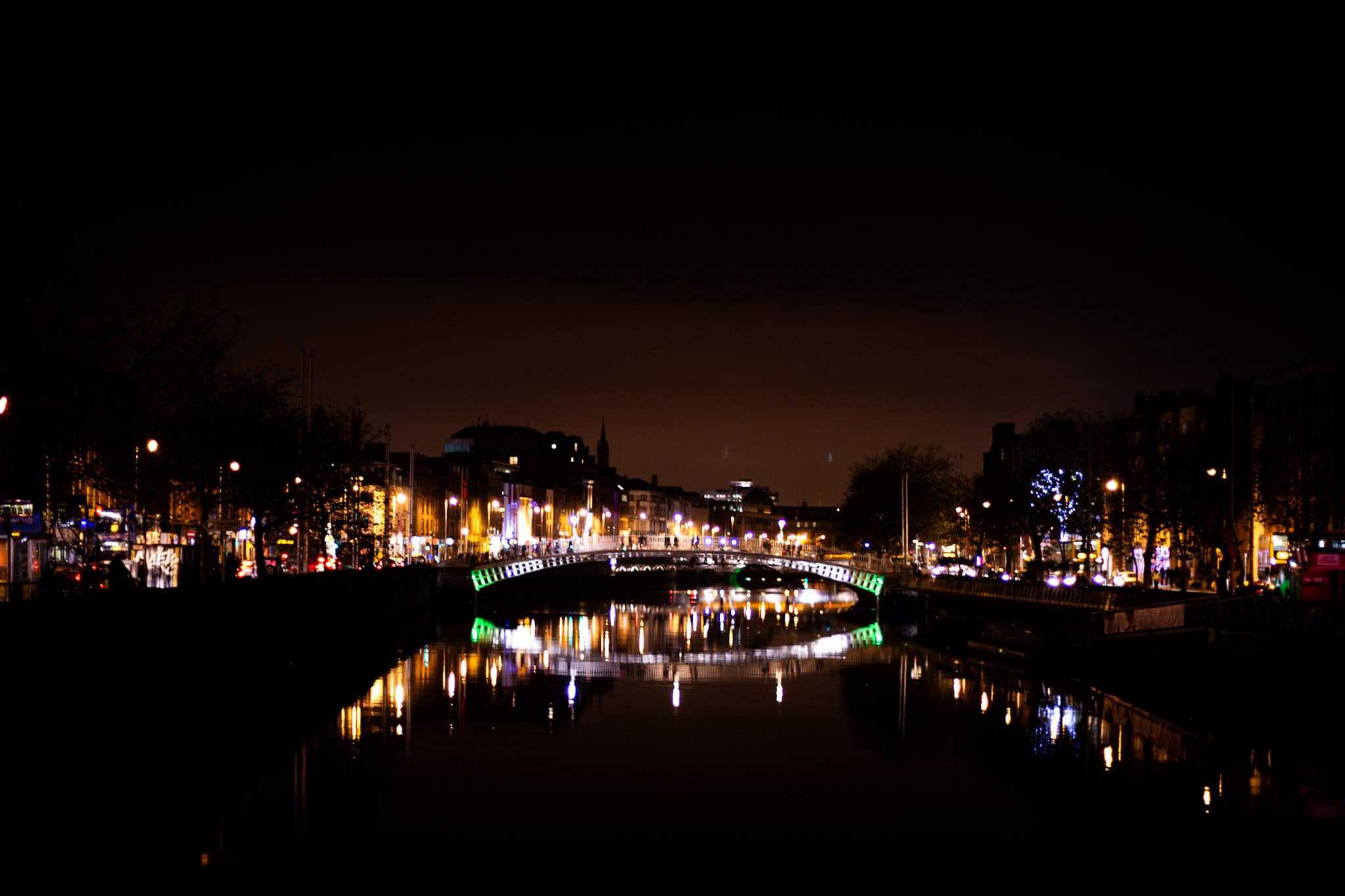 A wide-angle view of the Ha’penny Bridge lit in green and white, spanning the River Liffey. The bridge and surrounding city lights create a dazzling reflection on the calm water, framed by Dublin’s lively nighttime atmosphere.