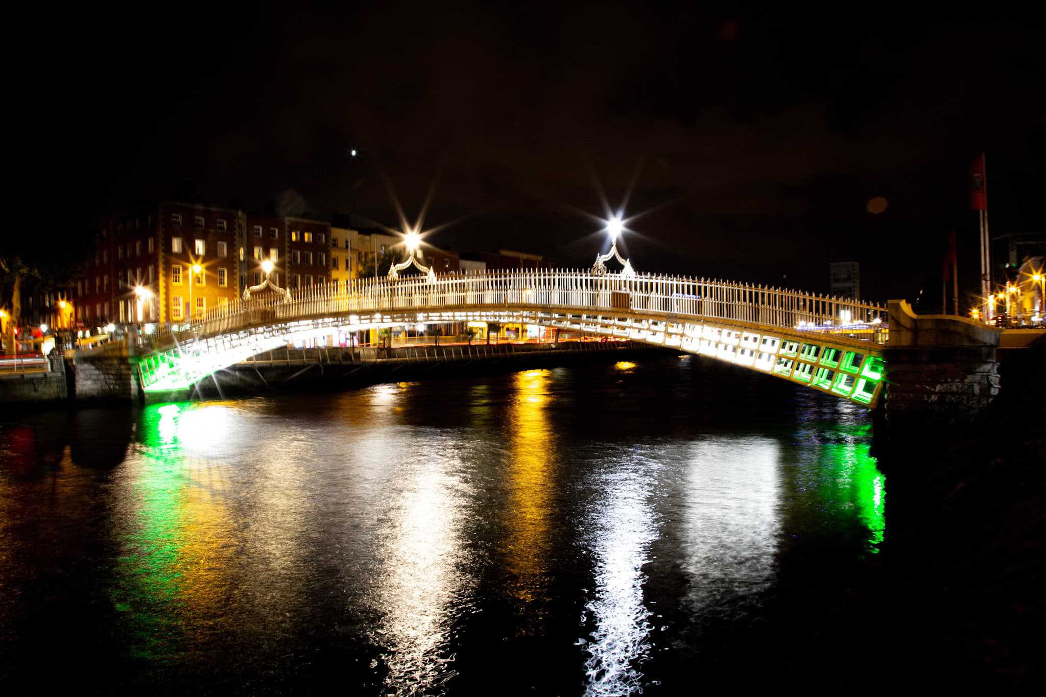 A nighttime view of Dublin’s iconic Ha’penny Bridge, illuminated with white and green lights reflecting in the River Liffey. The warm glow of nearby buildings enhances the picturesque scene under the dark sky.