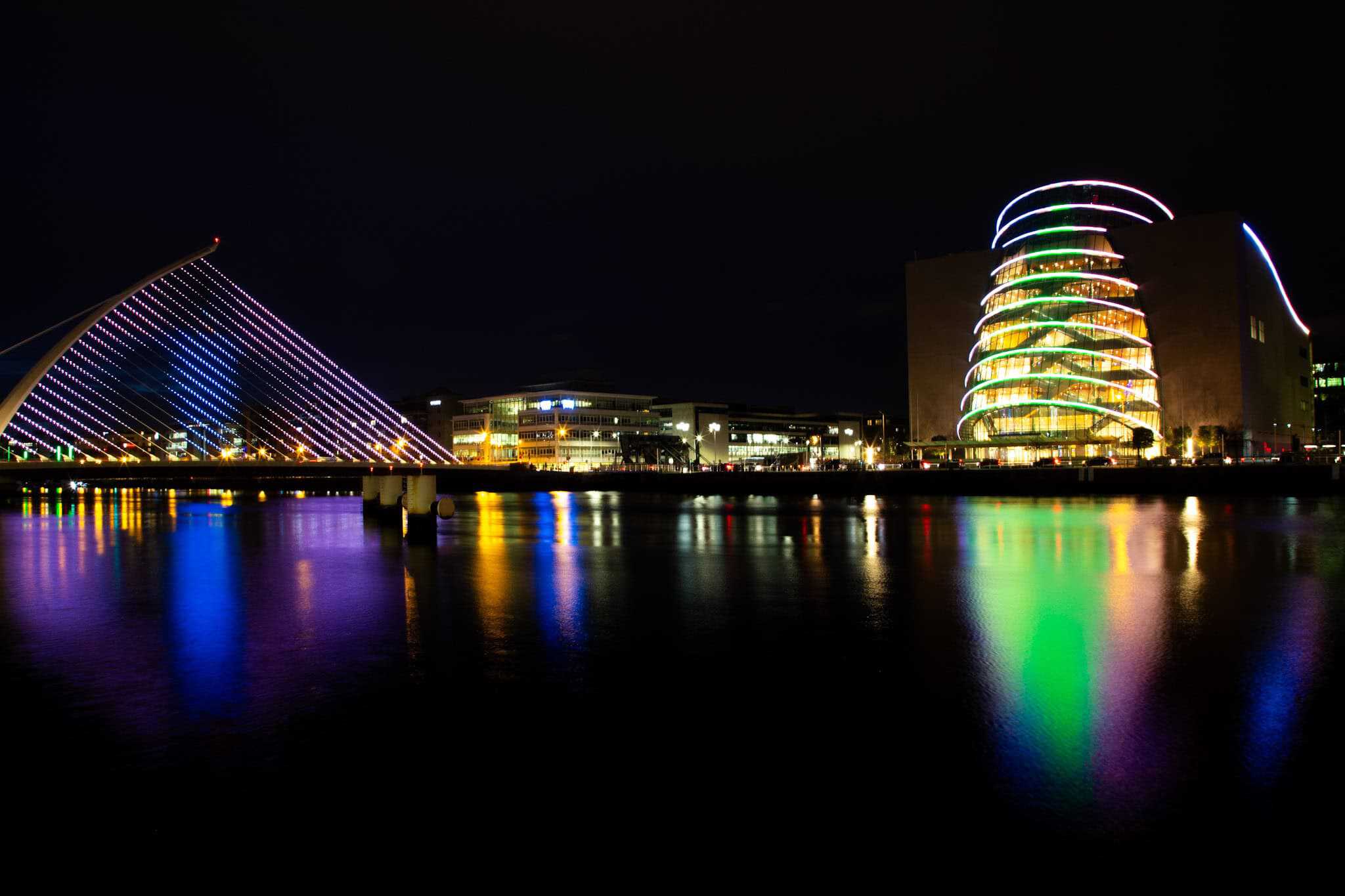 A night view of the Samuel Beckett Bridge, lit in vibrant pink hues, and the Dublin Convention Centre glowing with colorful stripes. The reflections shimmer across the dark waters of the River Liffey.