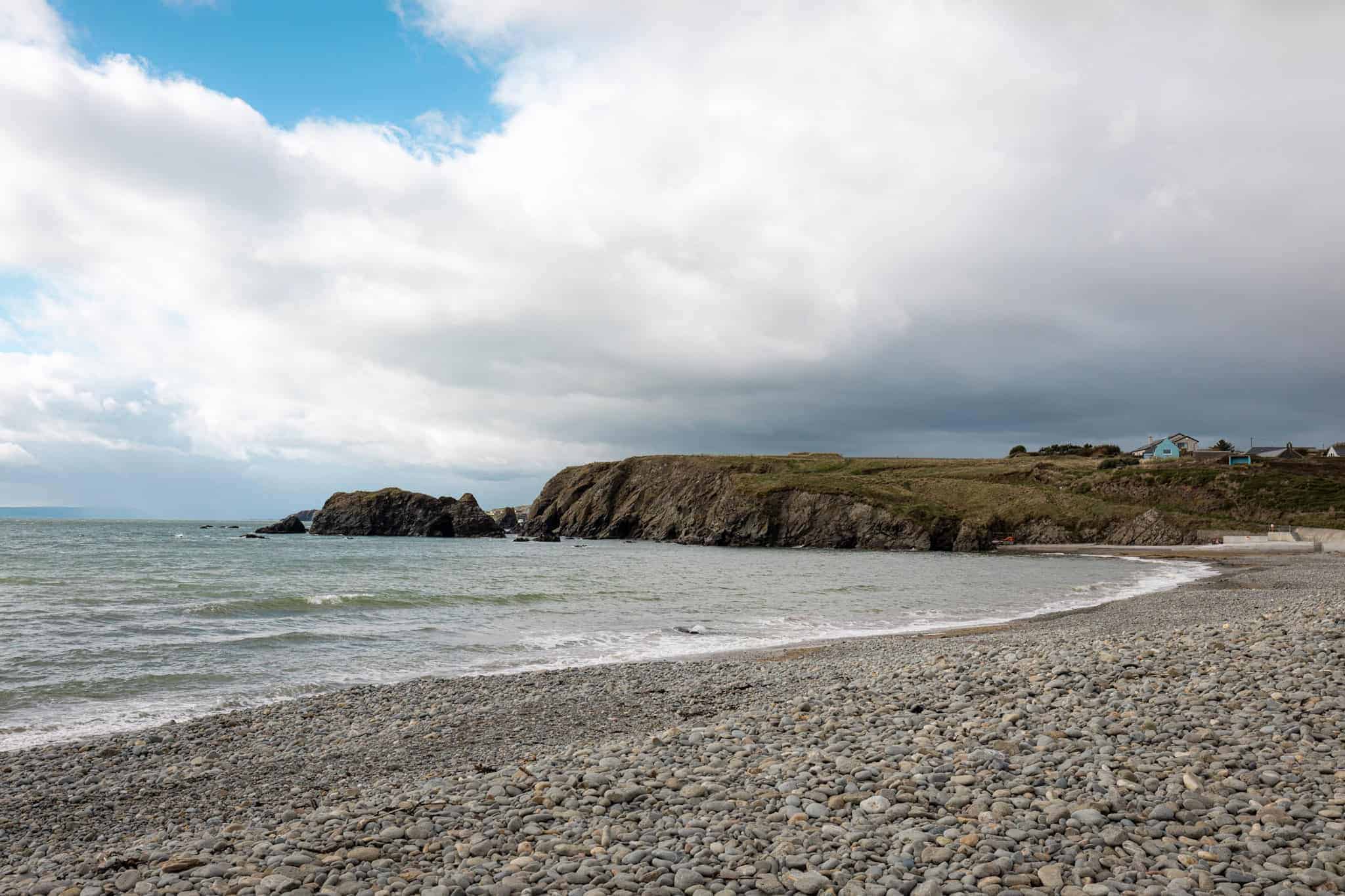 A pebbled beach along a rocky coastline, with gentle waves lapping at the shore under a partly cloudy sky. Green cliffs and a few homes can be seen in the background.