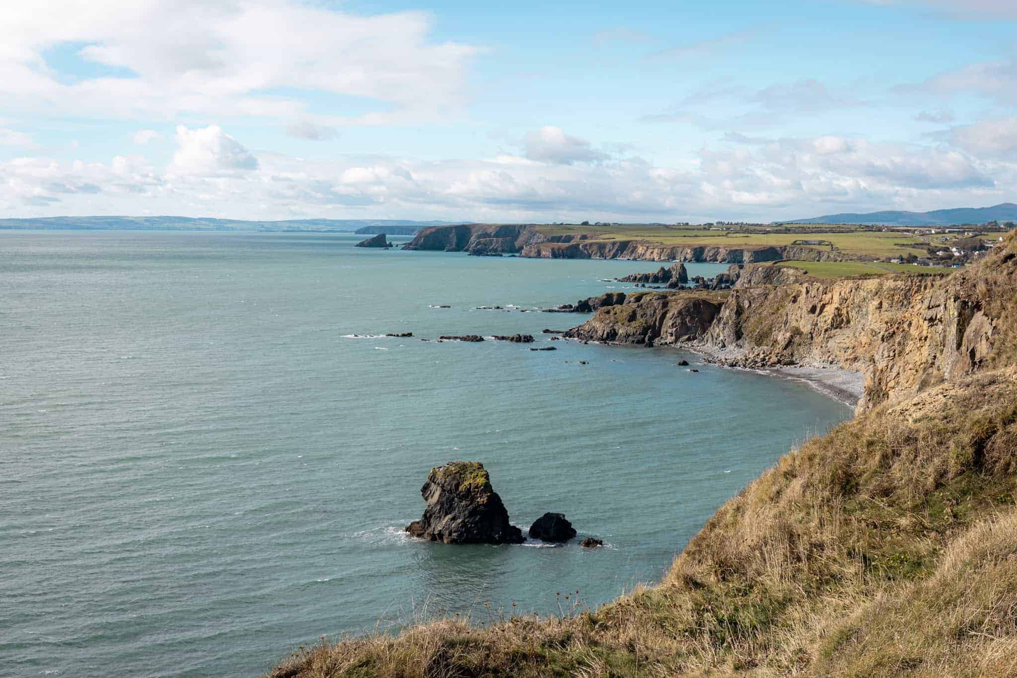 A dramatic view of rocky sea stacks rising from the water near steep cliffs. The ocean is a muted greenish-blue under a partly cloudy sky.