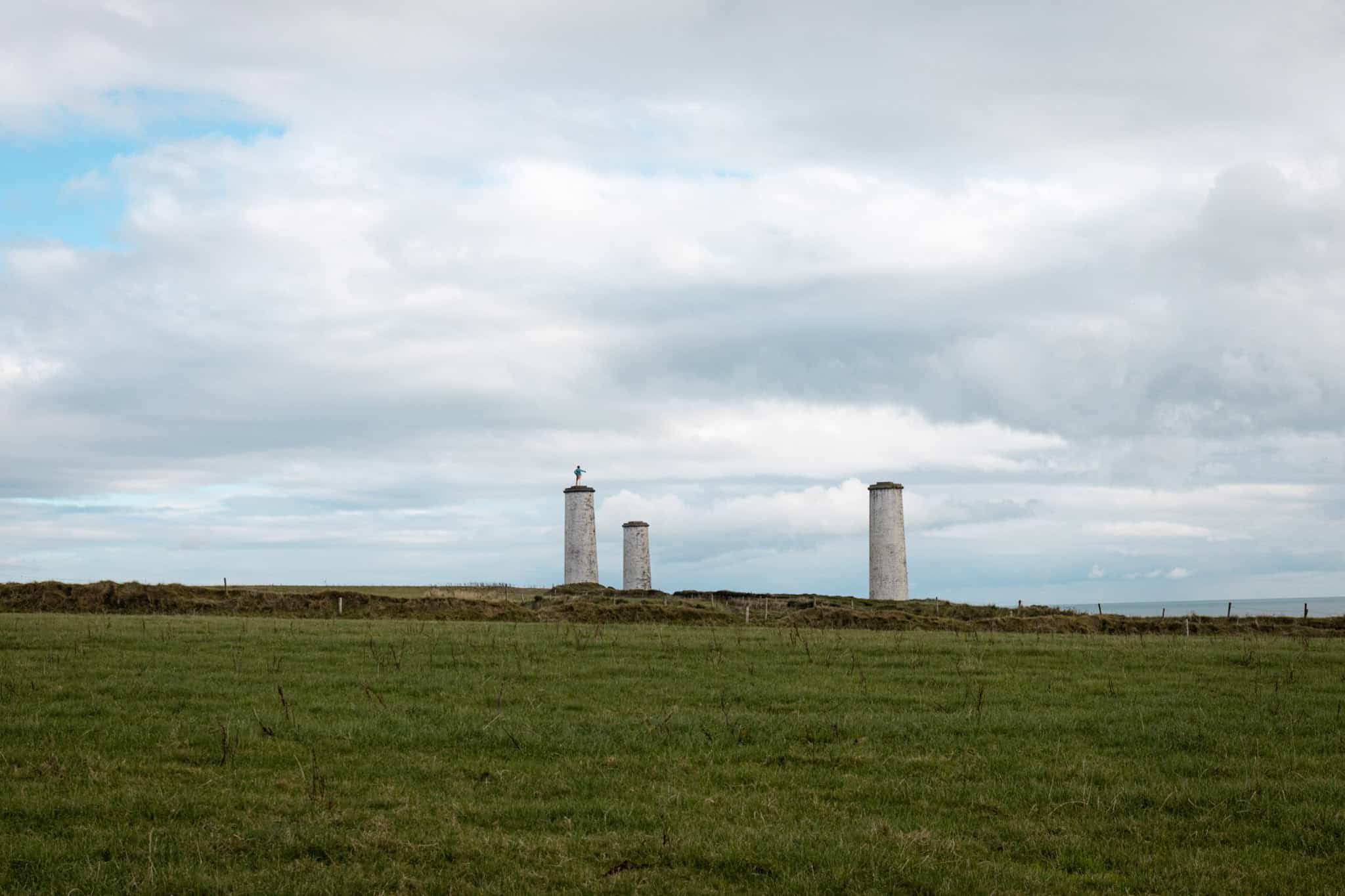 Three tall, cylindrical towers, one topped with a statue, stand on a grassy hill under a cloudy sky. The area is open and surrounded by rural landscape.