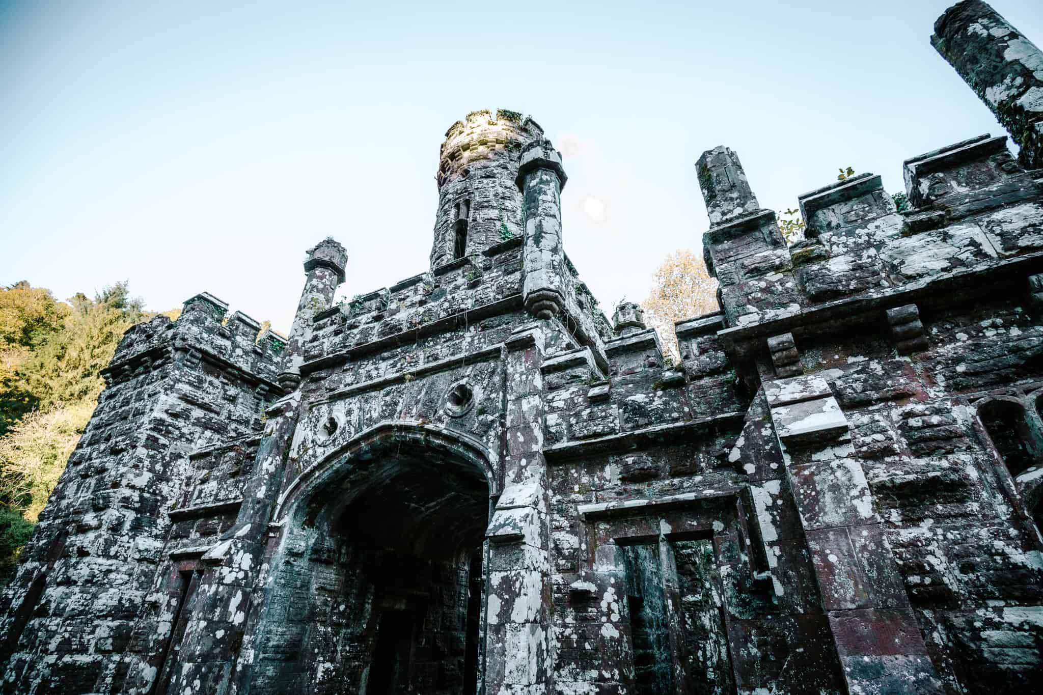 A towering, ancient stone gate of Ballysaggartmore Towers, partially covered in moss and lichen, with intricate Gothic architecture set against a bright, clear sky.