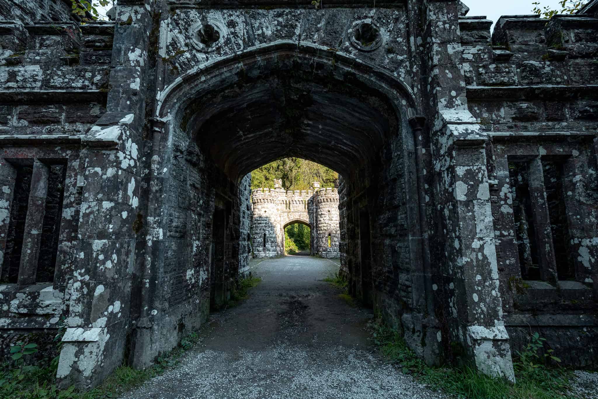 A close-up view of the dark, arched tunnel of Ballysaggartmore Towers, revealing a second arched tower further down the path, framed by rough stone walls.