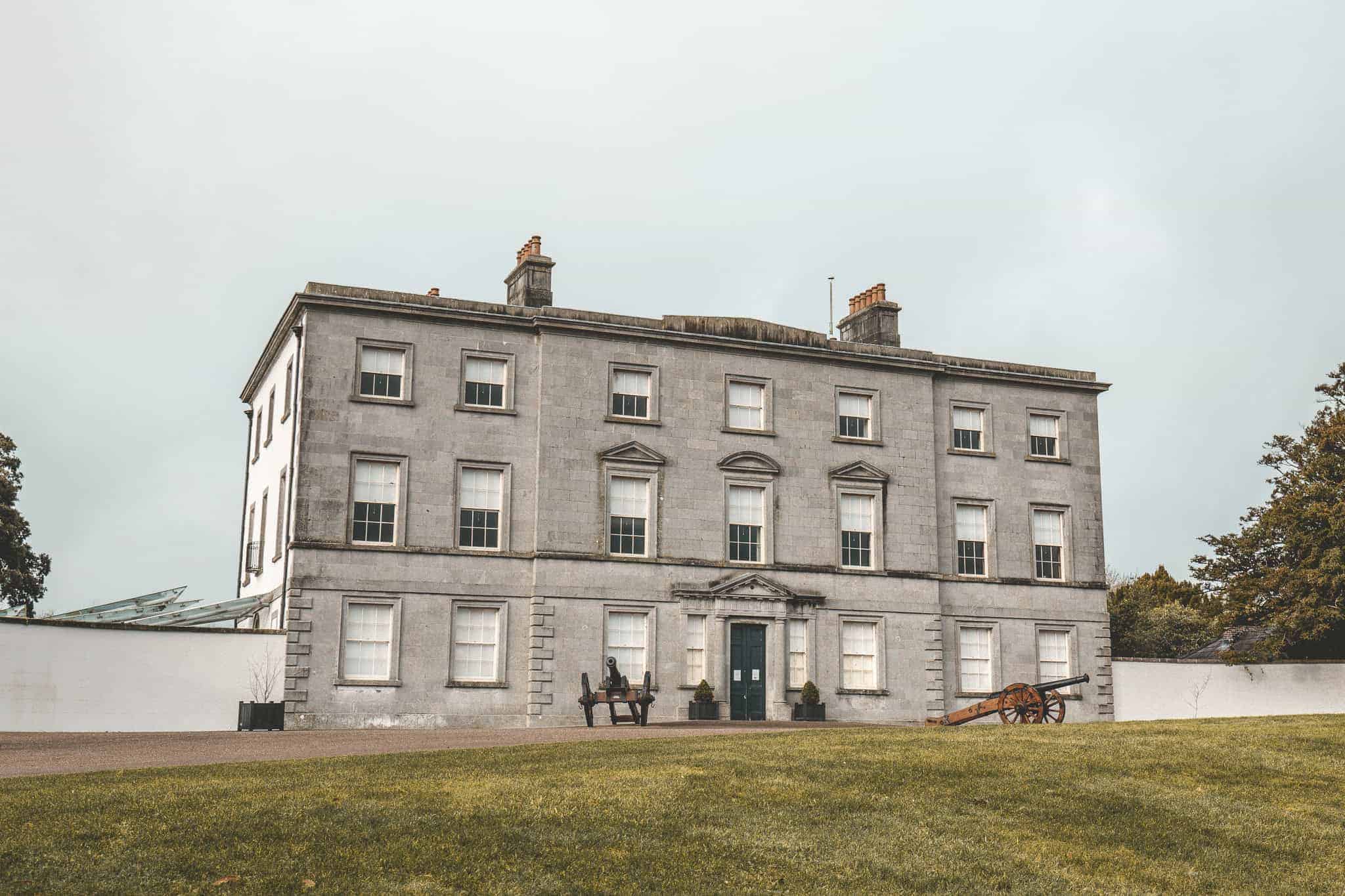grand historic building with a symmetrical facade and chimneys, surrounded by a grassy lawn. A cannon is displayed near the entrance, highlighting its historical significance as part of the Battle of the Boyne Visitor Center in Ireland.