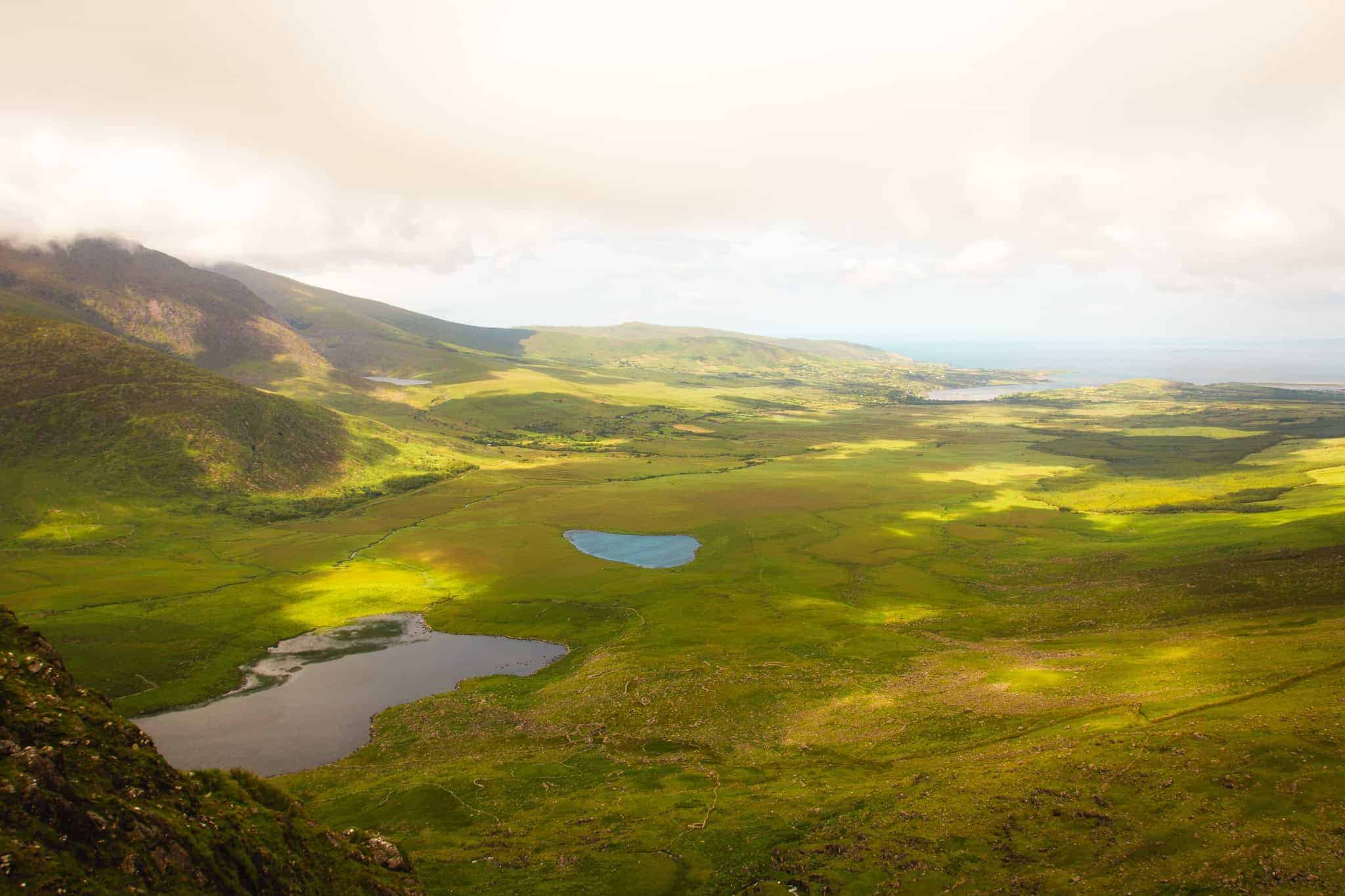 A scenic view of a lush green valley with two small lakes, rolling hills, and distant coastline under a hazy sky, illuminated by soft sunlight.