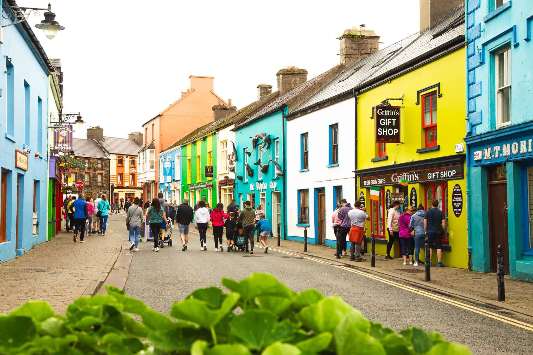 A lively street scene with people walking past vibrant, colorful buildings in shades of blue, green, yellow, and orange. One building has a sign for "Griffin's Gift Shop" selling Irish souvenirs.