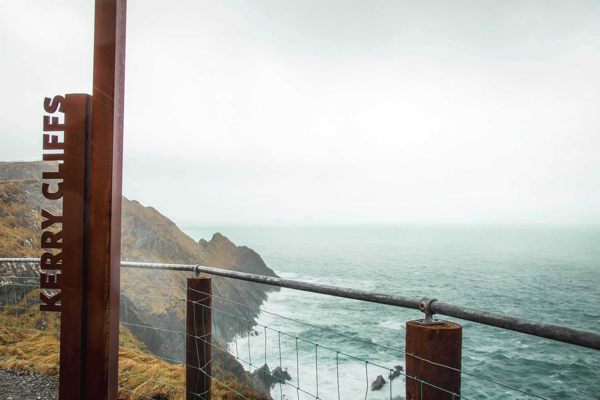 A dramatic view of the rugged Kerry Cliffs overlooking the Atlantic Ocean on a misty day. A metal railing and a tall post with "KERRY CLIFFS" cut into it are in the foreground.