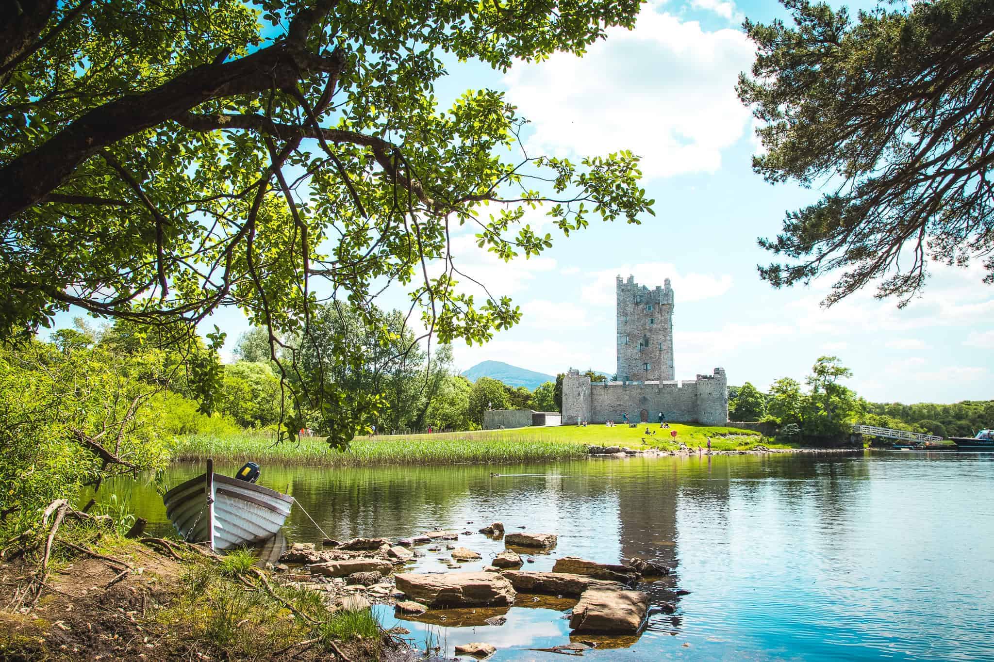 A peaceful lake scene with Ross Castle in the background, surrounded by trees. A small rowboat is tied to the shore in the foreground.