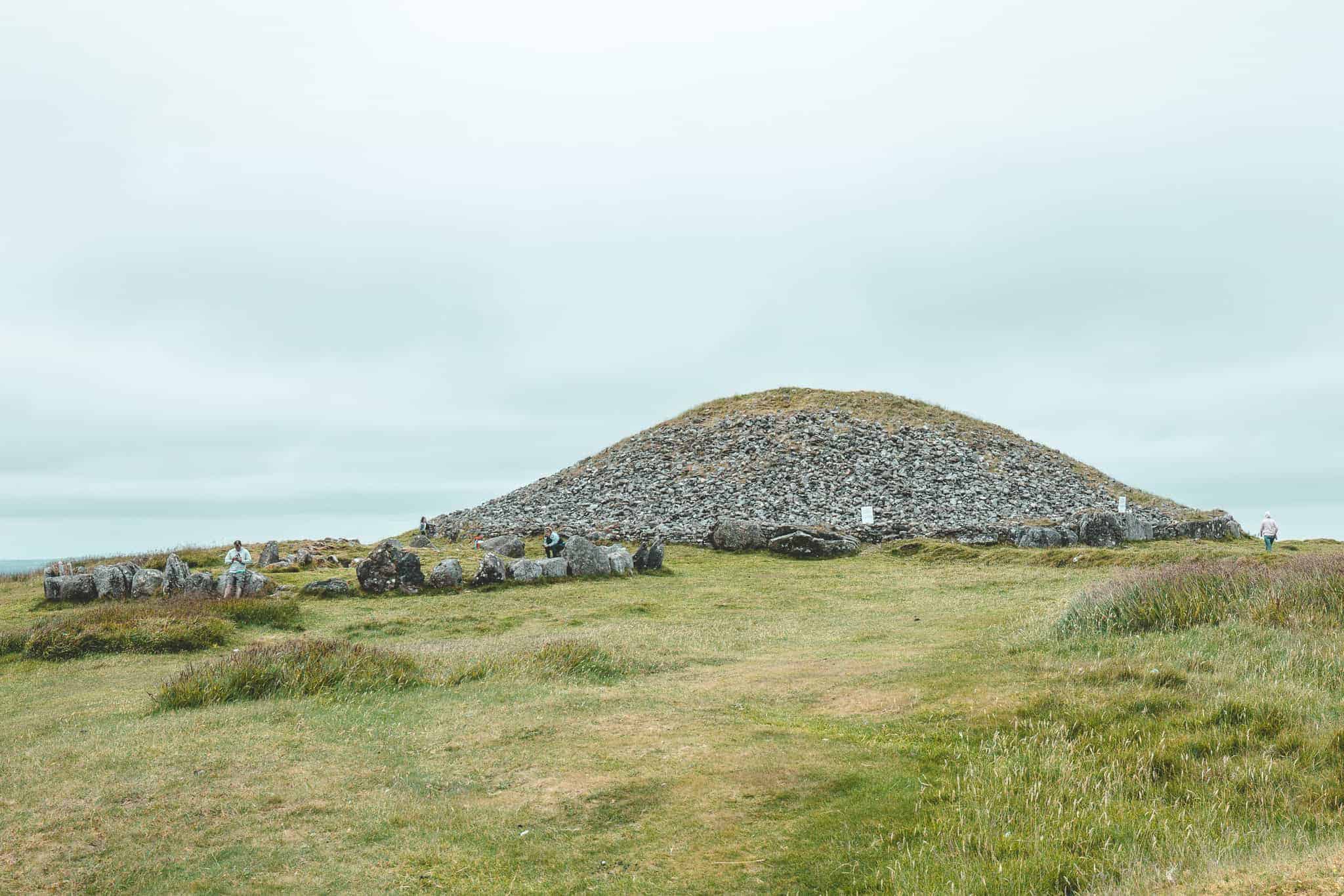 A large, grass-covered stone cairn at Loughcrew Cairns, with a circular mound made of piled rocks dominating the landscape. The foreground features scattered boulders and grassy terrain, while a few visitors are seen exploring the site under a cloudy sky.