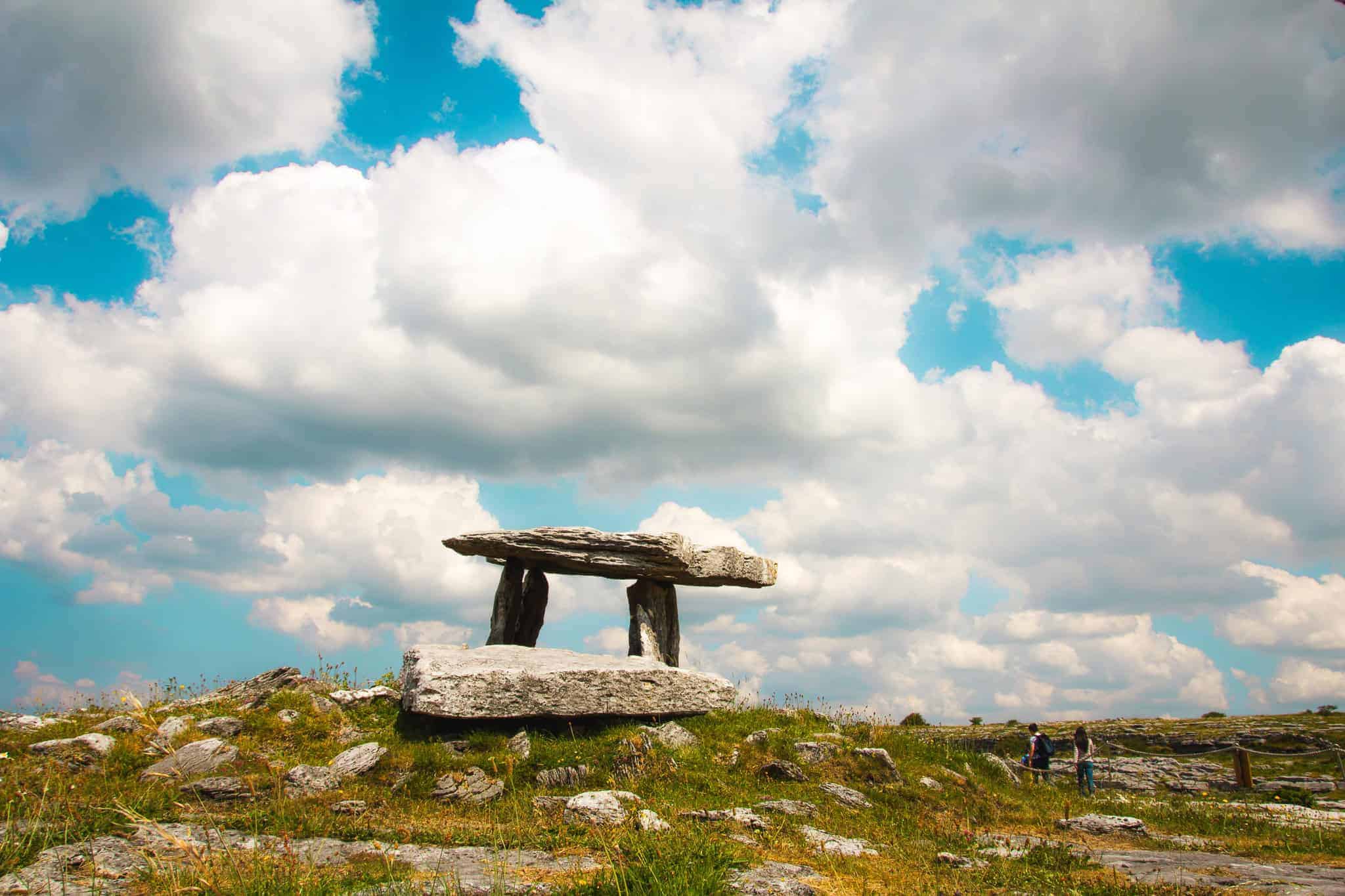 The ancient Poulnabrone Dolmen, a large stone portal tomb in The Burren, Ireland, stands under a blue sky with fluffy white clouds. Two people are seen exploring in the distance.